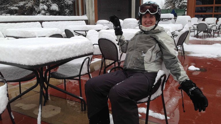 Diane Smith-Gander in ski gear sits in front of a table with several inches of snow on it