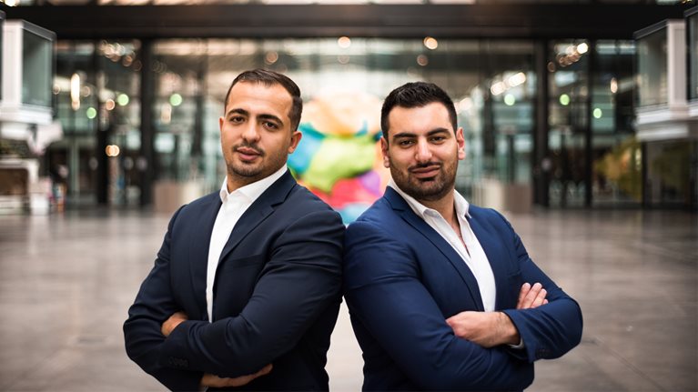 Amine Bounoughaz and his Edbridg co-founder both wearing blue suits stand back to back facing camera and smiling
