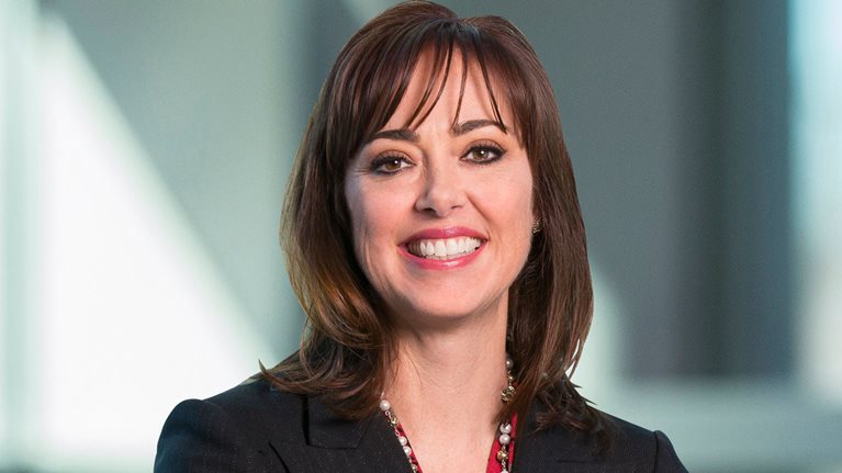 Monica McGurk smiling in front of a gray background wearing a black dress