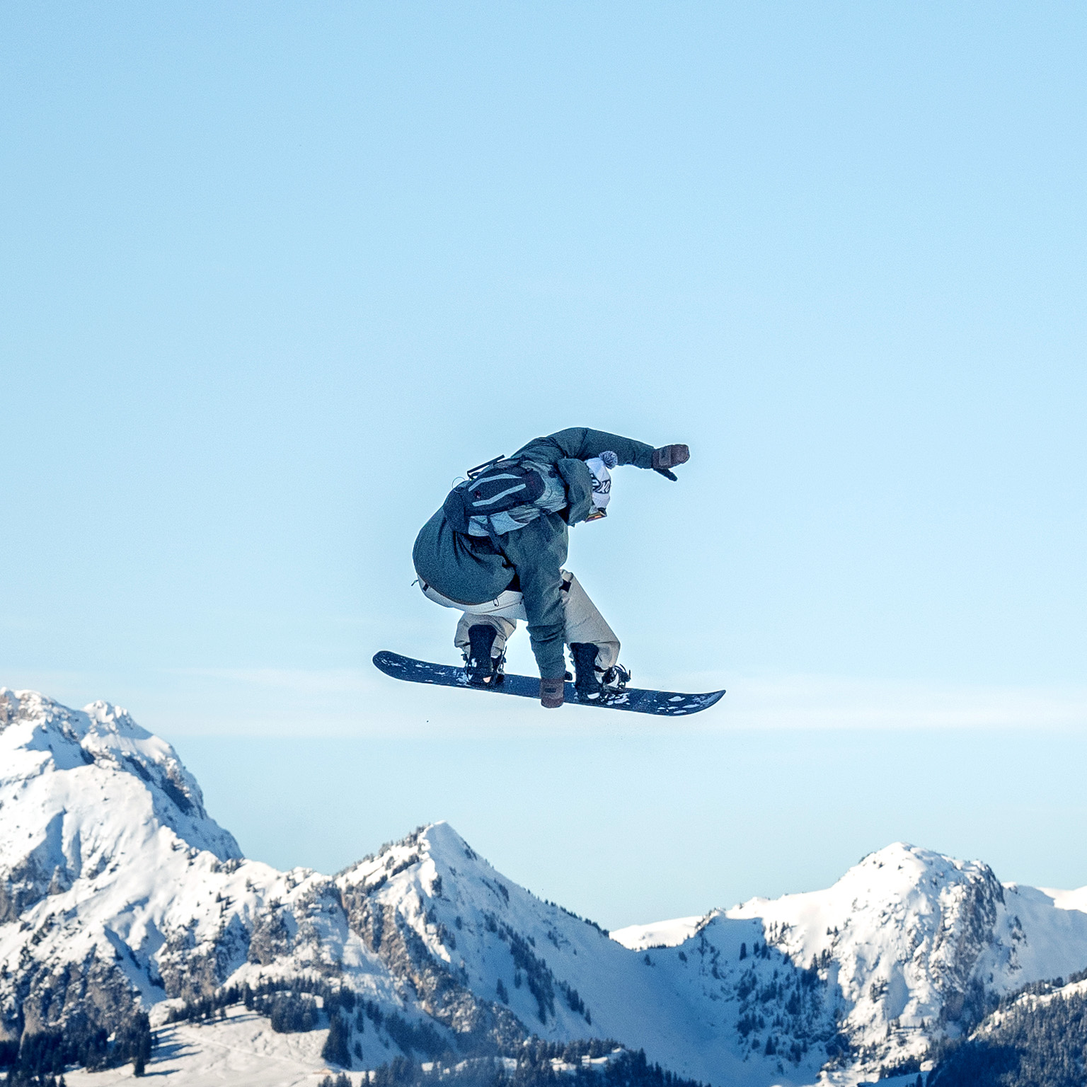 A snowboarder is captured midair performing a jump against a clear blue sky, with rugged, snow-covered mountains stretching across the background. The scene conveys motion and adventure, highlighting the contrast between the airborne rider and the vast alpine landscape below.