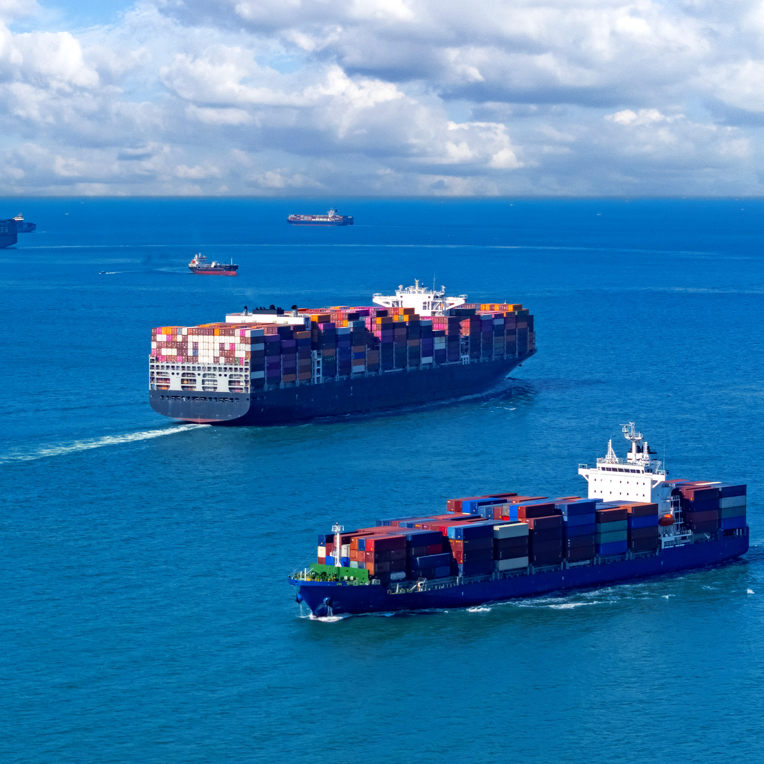 An image of several container ships navigating a vast expanse of blue ocean under a sky filled with fluffy white clouds.