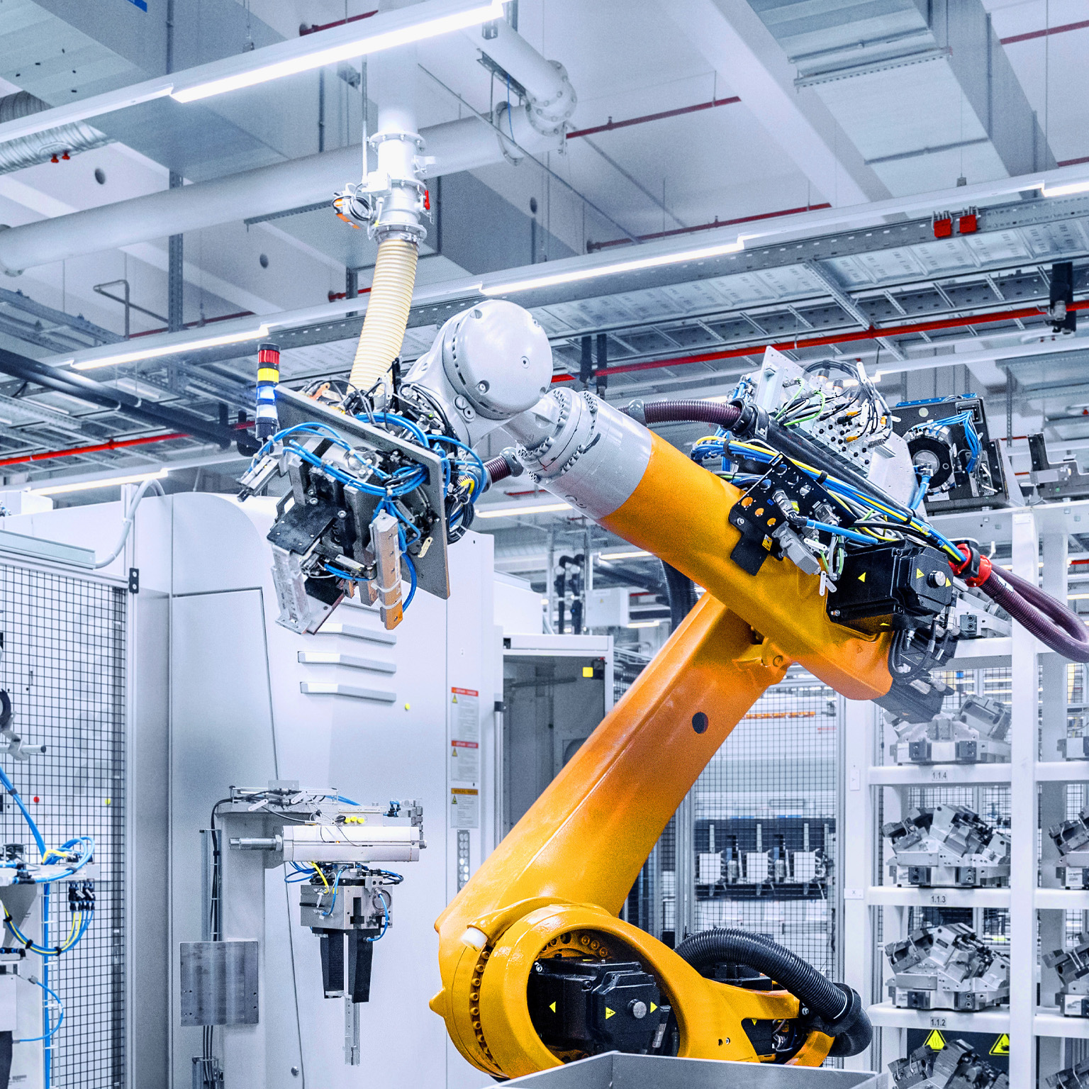 A large, bright industrial workspace with a prominent yellow robotic arm in the foreground. The robot appears to be working within an automated manufacturing system, surrounded by other machinery, parts, and storage racks.