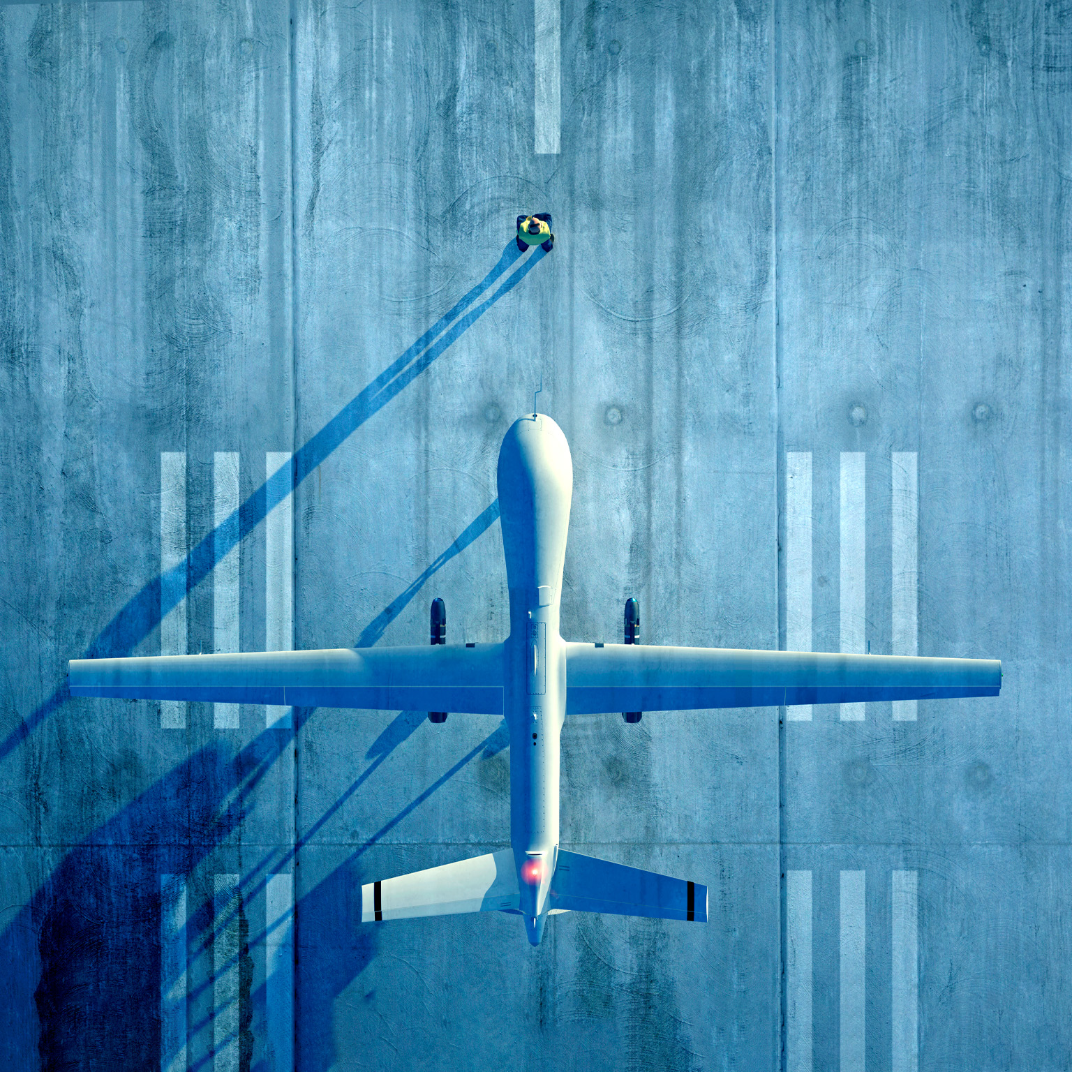 An overhead view shows a large unmanned aircraft parked on a concrete runway, its long wings stretching symmetrically across the frame. A small person nearby casts a long shadow, emphasizing the scale of the scene.