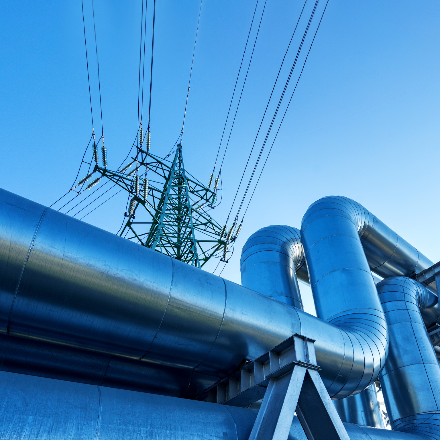 Large metal pipes tansporting oil curve and ascend against a clear blue sky with a tall electricity pylon visible in the background.
