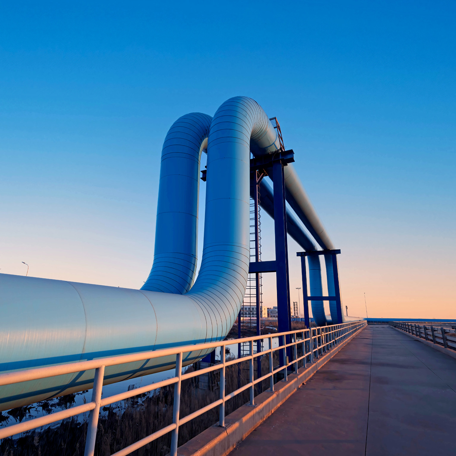 A large industrial pipeline rises and bends over a bridge-like structure, set against a clear blue sky at dusk. A paved walkway with railings runs alongside the pipes, leading the eye toward the horizon and giving the scene a clean, modern, and slightly futuristic feel.