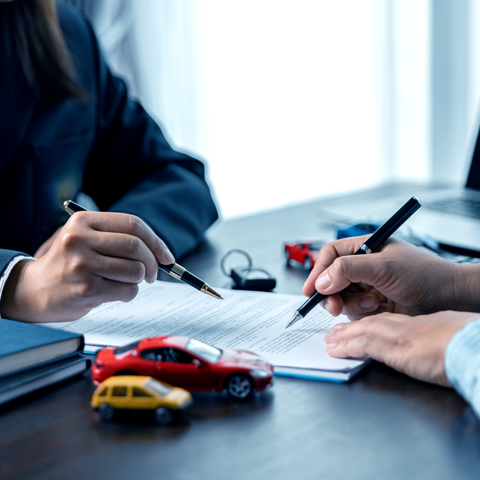 Two people signing documents at a desk, with their hands holding pens over a contract. Toy cars and a set of keys on the table suggest the paperwork is related to an auto insurance agreement.