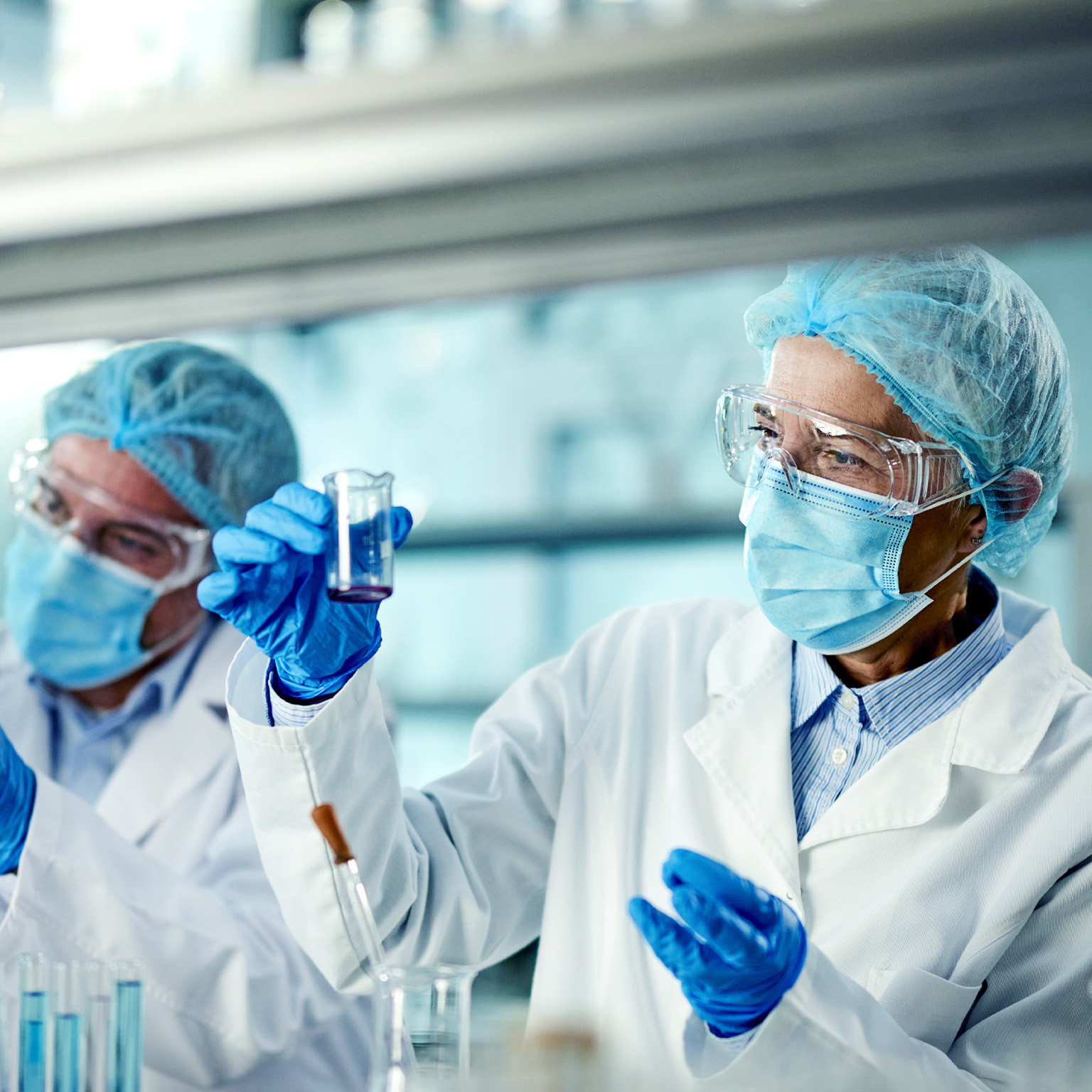 Two scientists in a laboratory are wearing protective gear including lab coats gloves and face masks One scientist carefully examines a liquid in a beaker while the other holds test tubes containing samples.