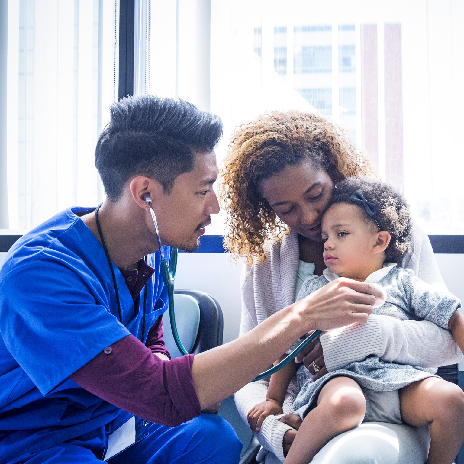 A young doctor in a blue scrub is using a stethoscope to examine a toddler who is sitting on their mother's lap.