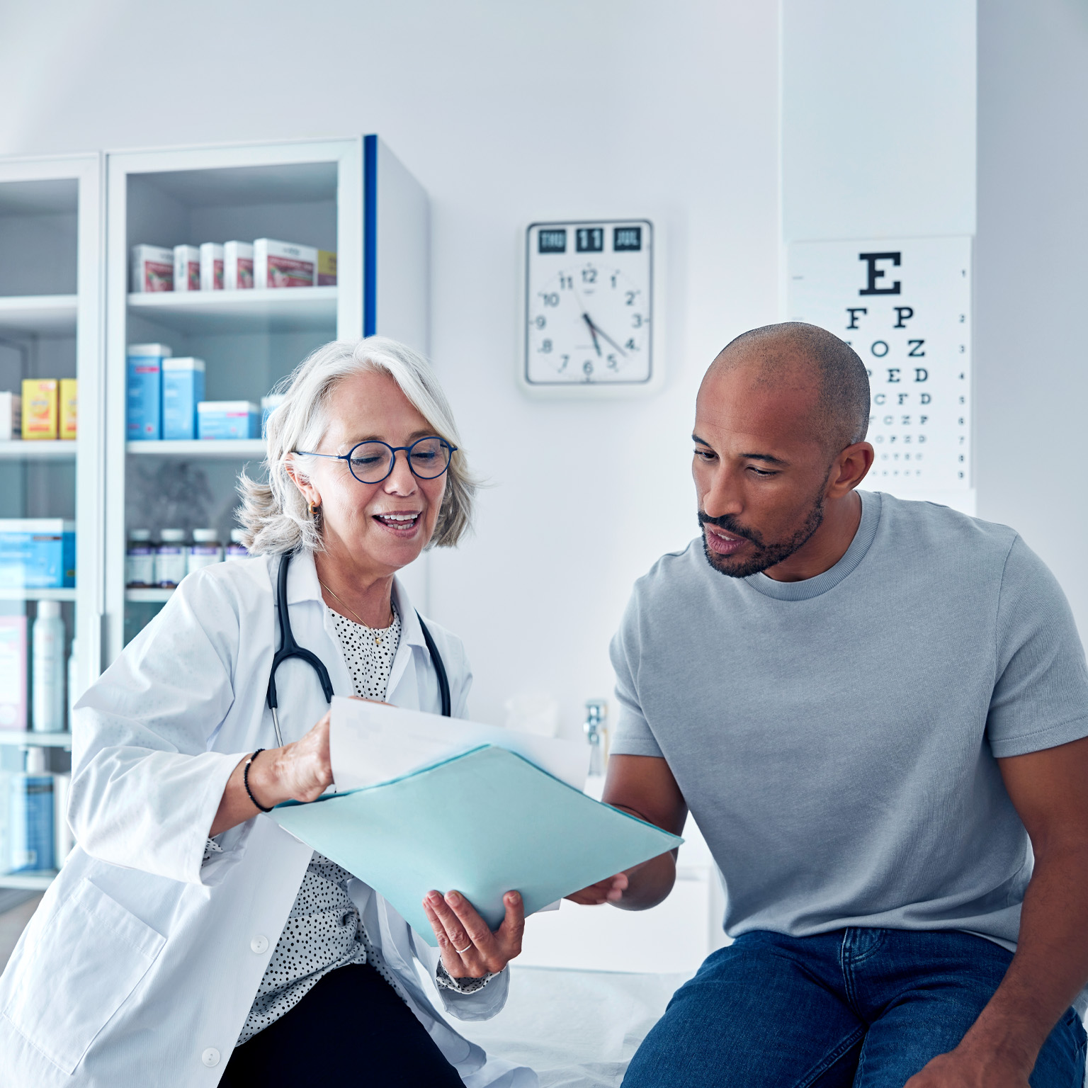 A doctor and a patient sitting together in a medical exam room, reviewing documents and discussing information in a folder. The setting includes medical cabinets, charts, and equipment, creating a calm, professional healthcare environment.