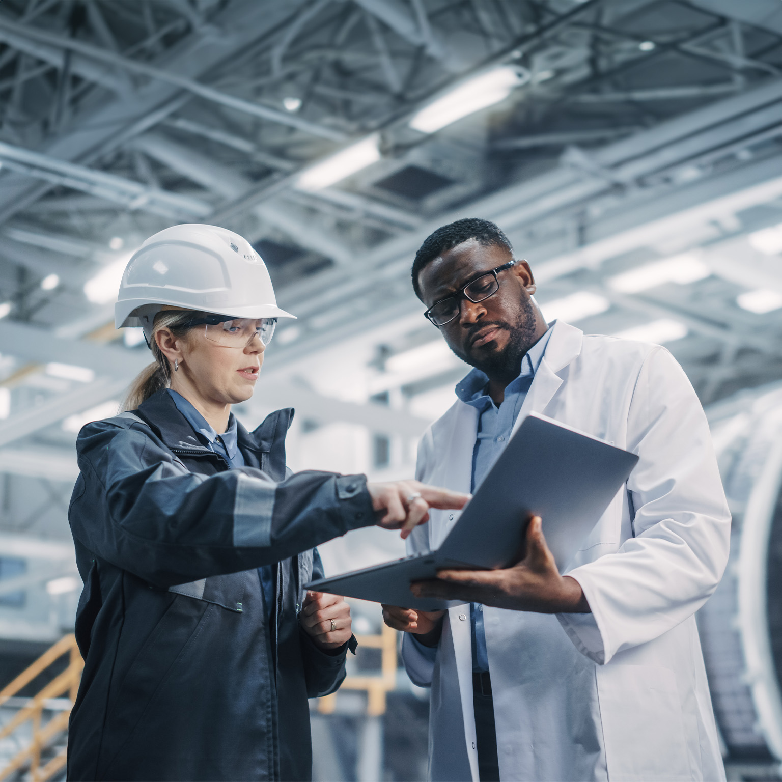 Team of Diverse Professional Heavy Industry Engineers Wearing Safety Uniform and Hard Hat Working on Laptop Computer. African American Technician and Female Worker Talking on a Meeting in a Factory. - stock photo