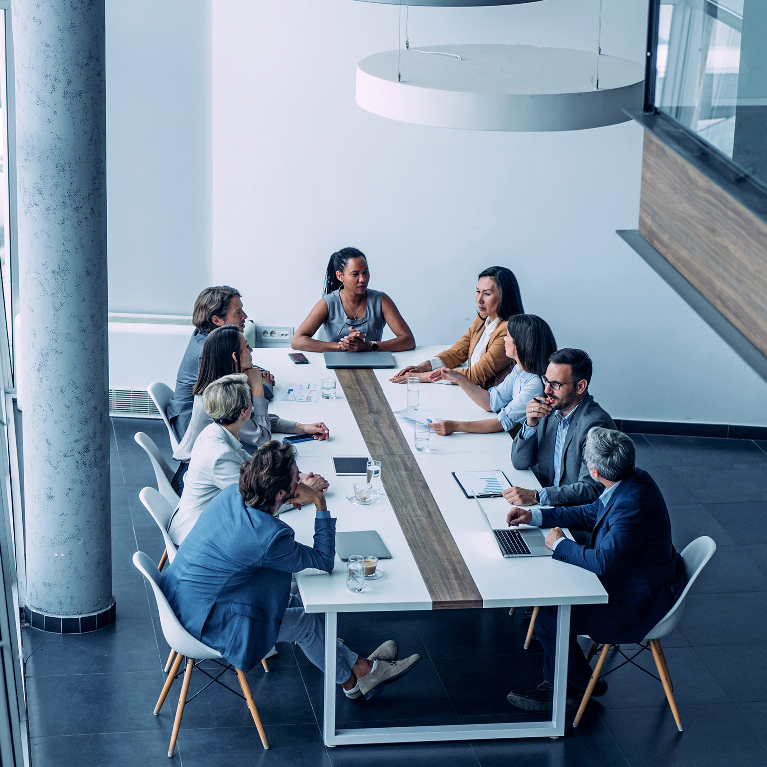 In a modern office setting with bright lighting and large windows, a diverse group of professionals gathers around a long conference table, seemingly engaged in a business meeting.