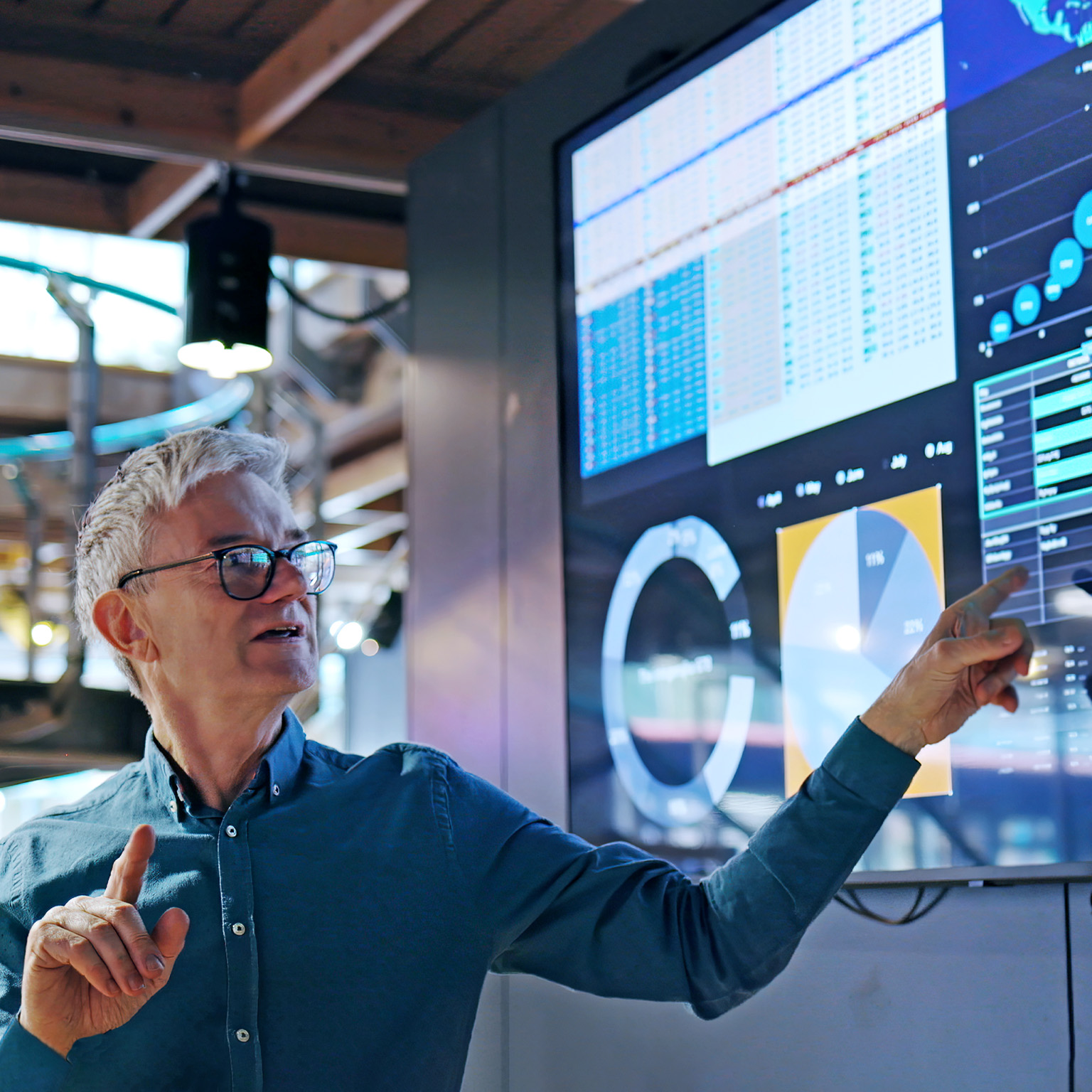 A man gesturing towards a large screen displaying graphs and data as he leads a presentation.