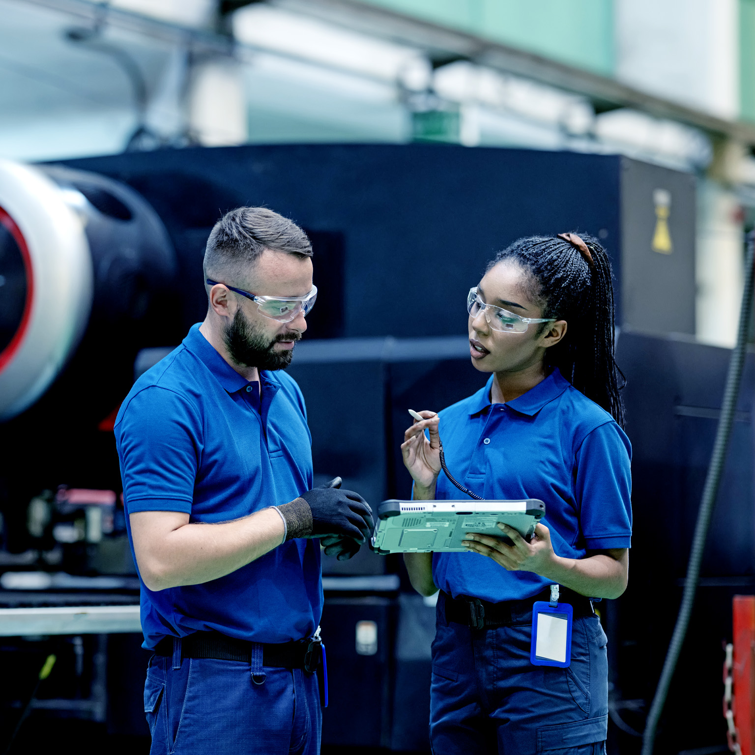 In an industrial manufacturing setting, a supervisor and process engineer, both dressed in blue company shirts and wearing protective glasses, engage in a conversation while examining a tablet near a sizable machine.