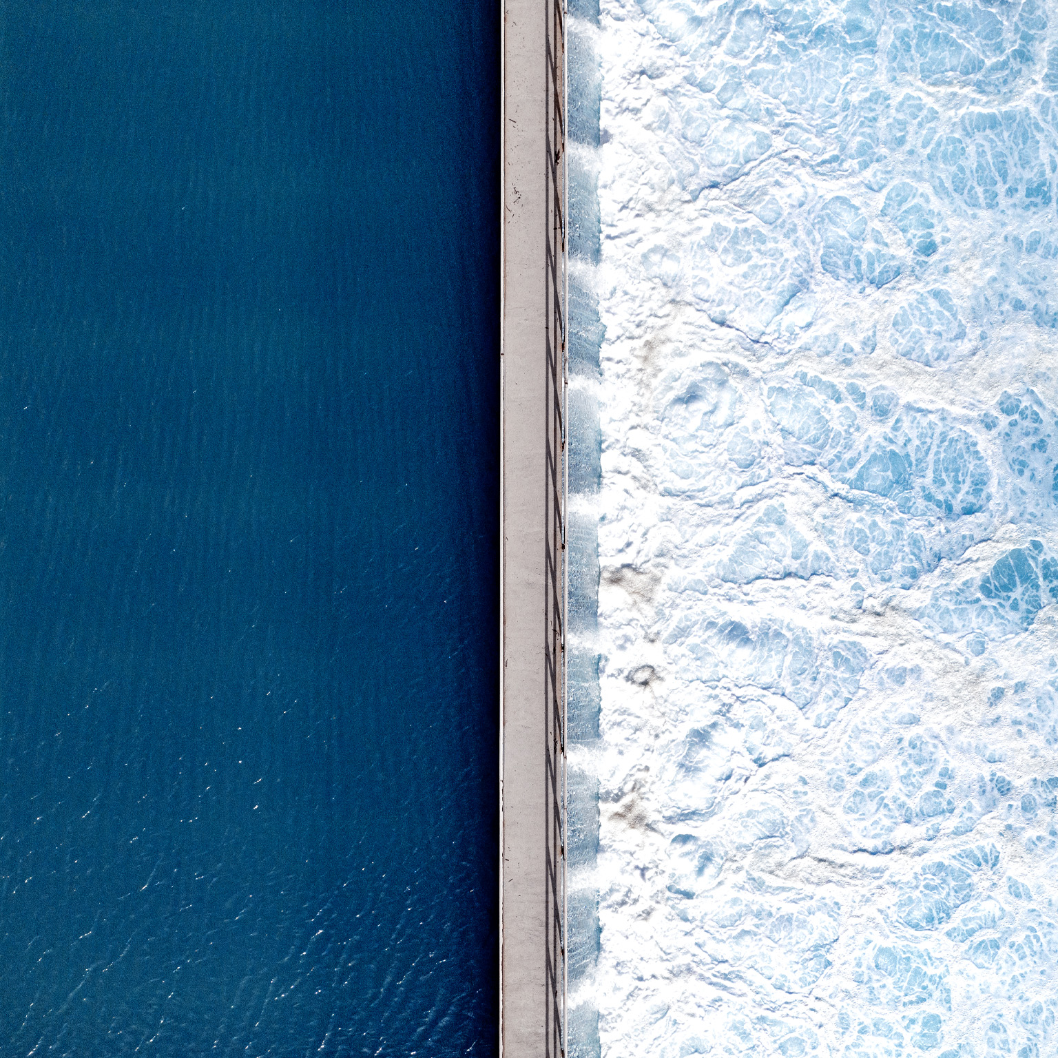 An overhead shot captures the stark contrast between deep, tranquil blue water on one side and churning, white-foamed turquoise waves on the other, bisected by a narrow concrete pier. The smooth, dark surface on the left stands in dramatic opposition to the textured, frothy turbulence crashing against the right side of the man-made barrier.