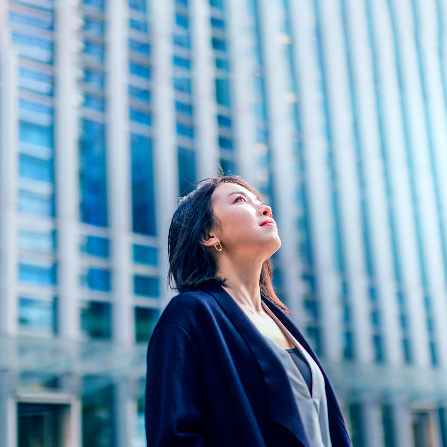 A modern business district with tall buildings is the background for a young, confident businesswoman looking up at the sky from a low angle.