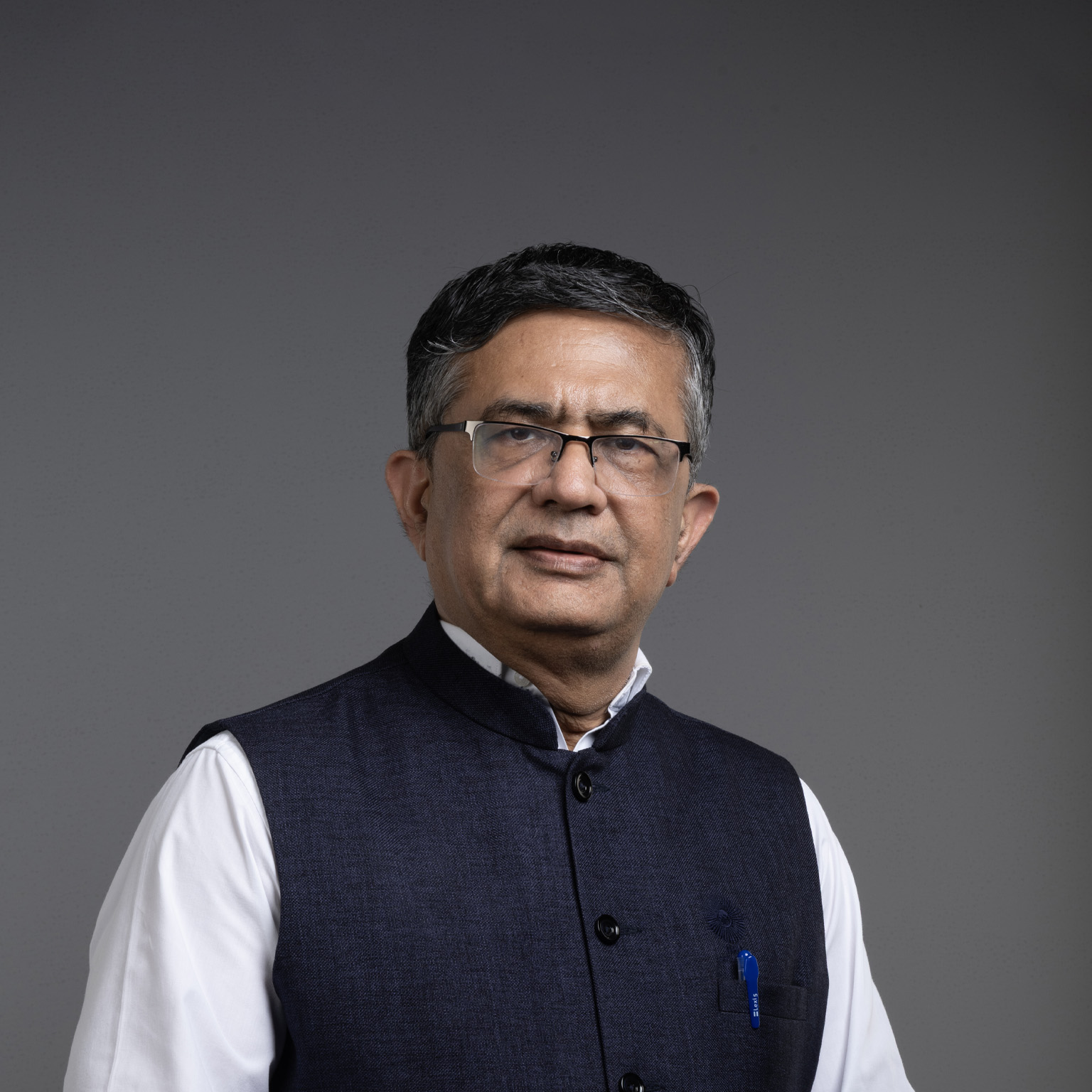 Ashishkumar Chauhan, with short hair and glasses, is wearing a white shirt and a dark blue Nehru vest, standing against a gray background.
