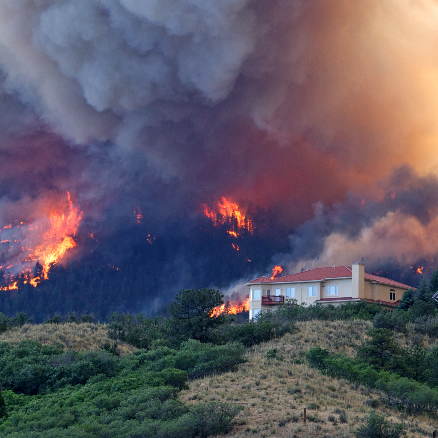 Image of a forest fire burning on a hillside, threatening houses below the blaze, with thick smoke and flames swirling and reaching for the sky.