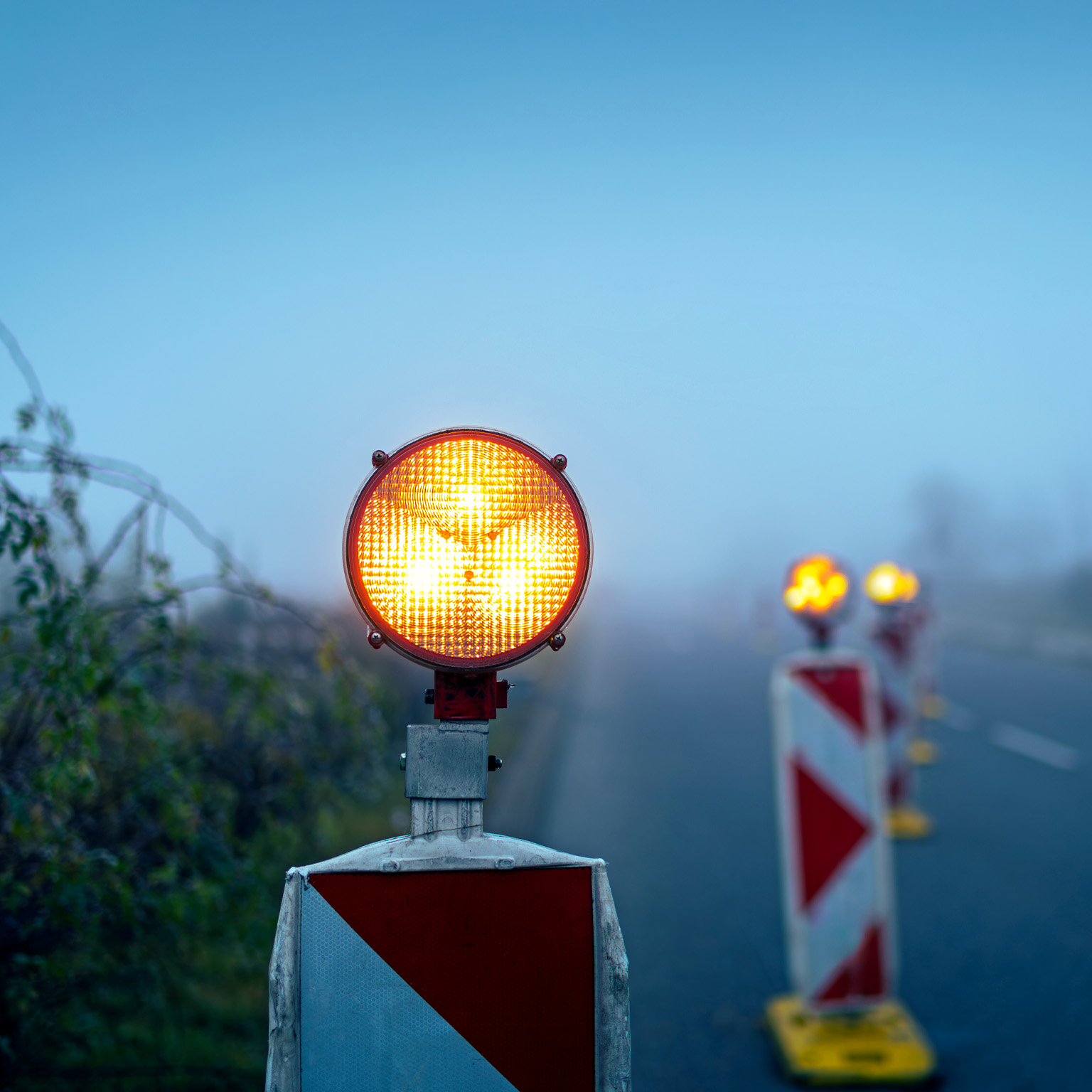 A bright amber warning light sits atop a road barrier, glowing vividly against a foggy, muted background. Additional blurred warning lights and striped barriers line the roadside.