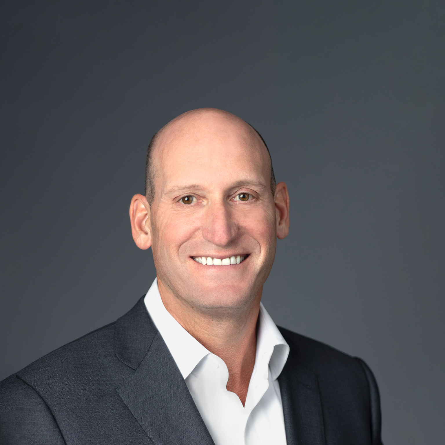 David Gitlin is standing against a neutral gray background, dressed in a dark suit jacket and white shirt, with a smile on his face.