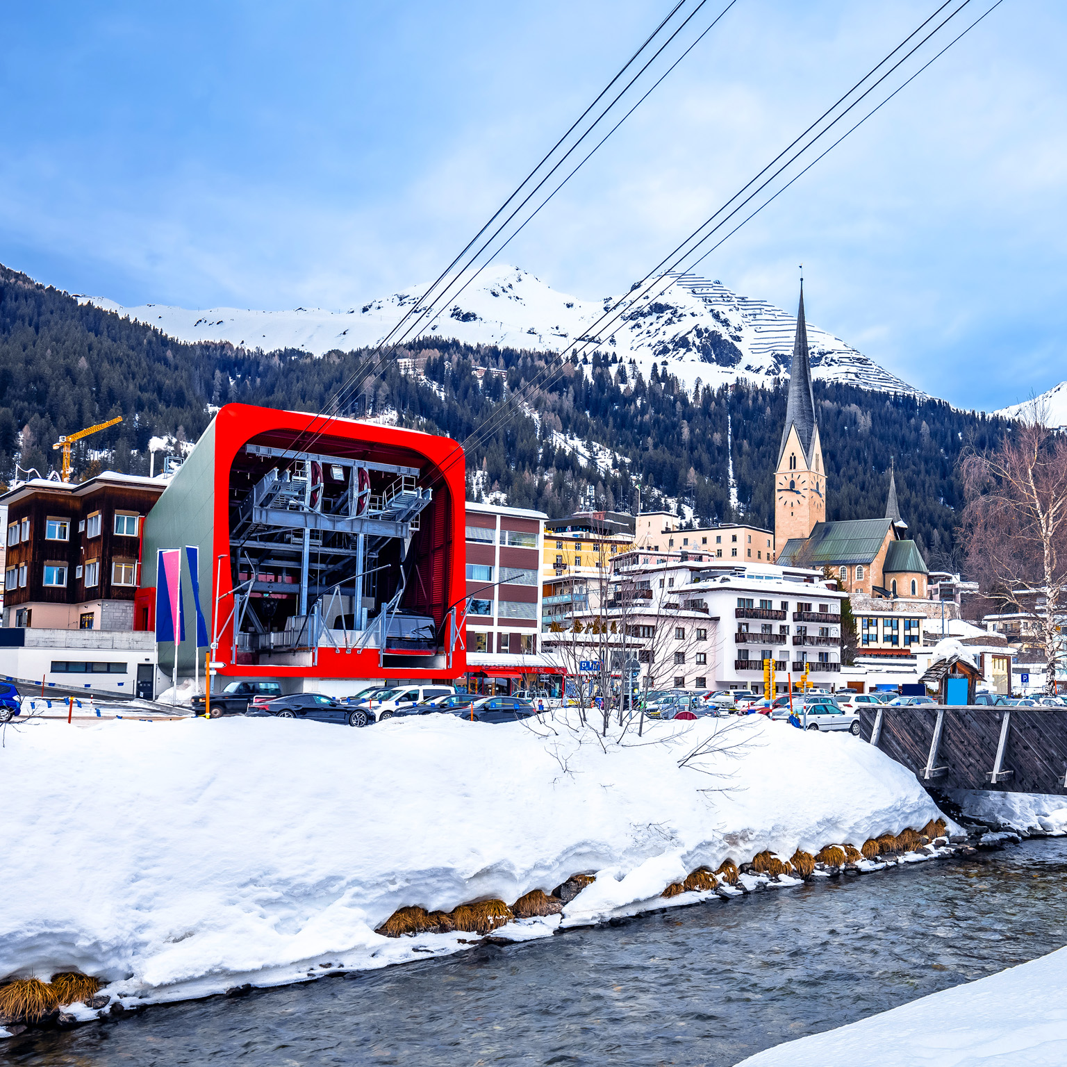 From Davos, Switzerland, one can see the tramway that takes you to the ski resort. Alongside the tram, there are small buildings, while the background is dominated by mountains. A stream flows in the foreground, adding to the overall view.