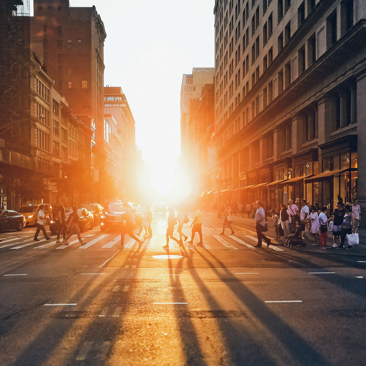 Sunlight shining on the diverse crowds of people walking across the busy intersection on 5th Avenue in New York City.