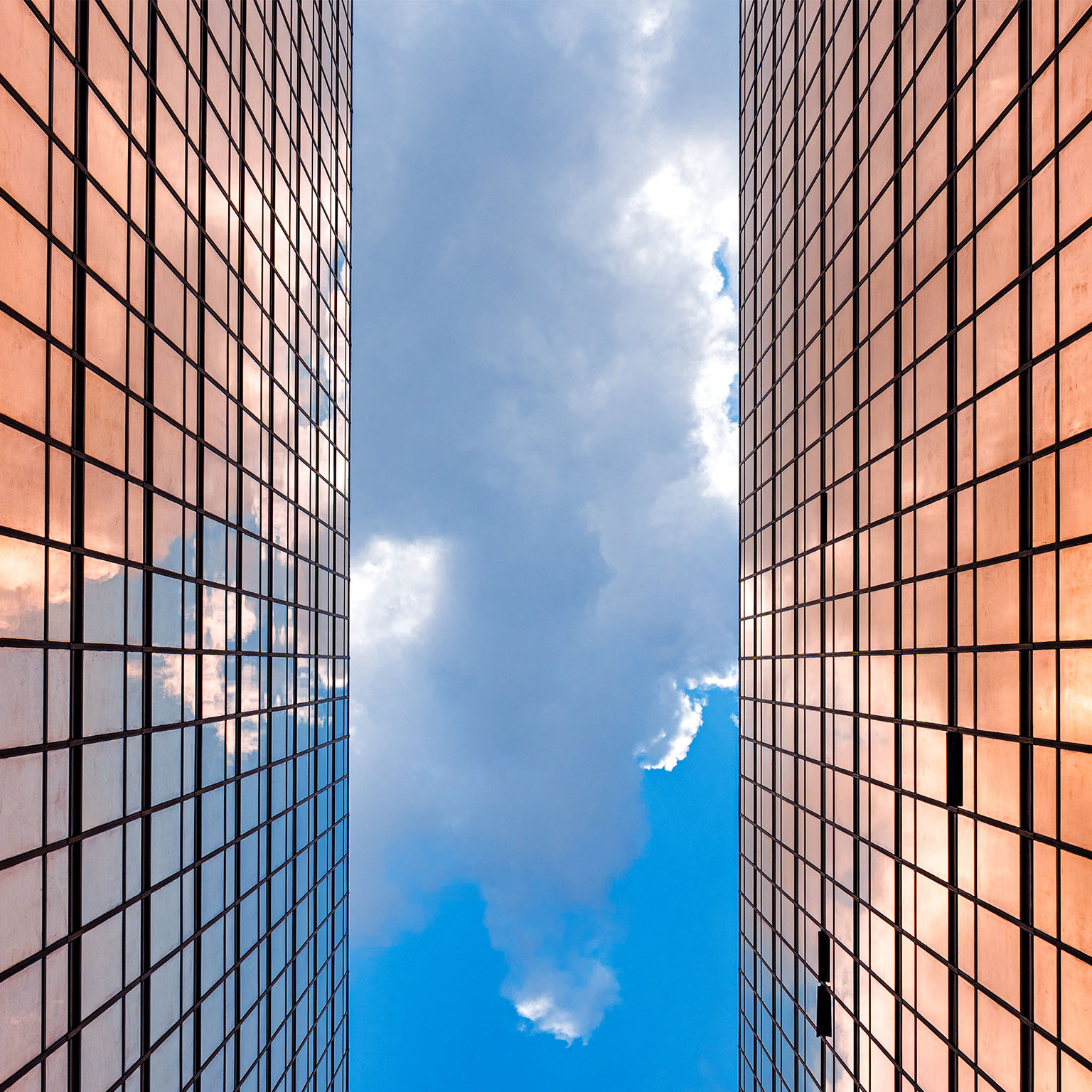 Looking up between two gleaming glass towers toward a vivid blue sky, the city reflects a bright future rising ahead.