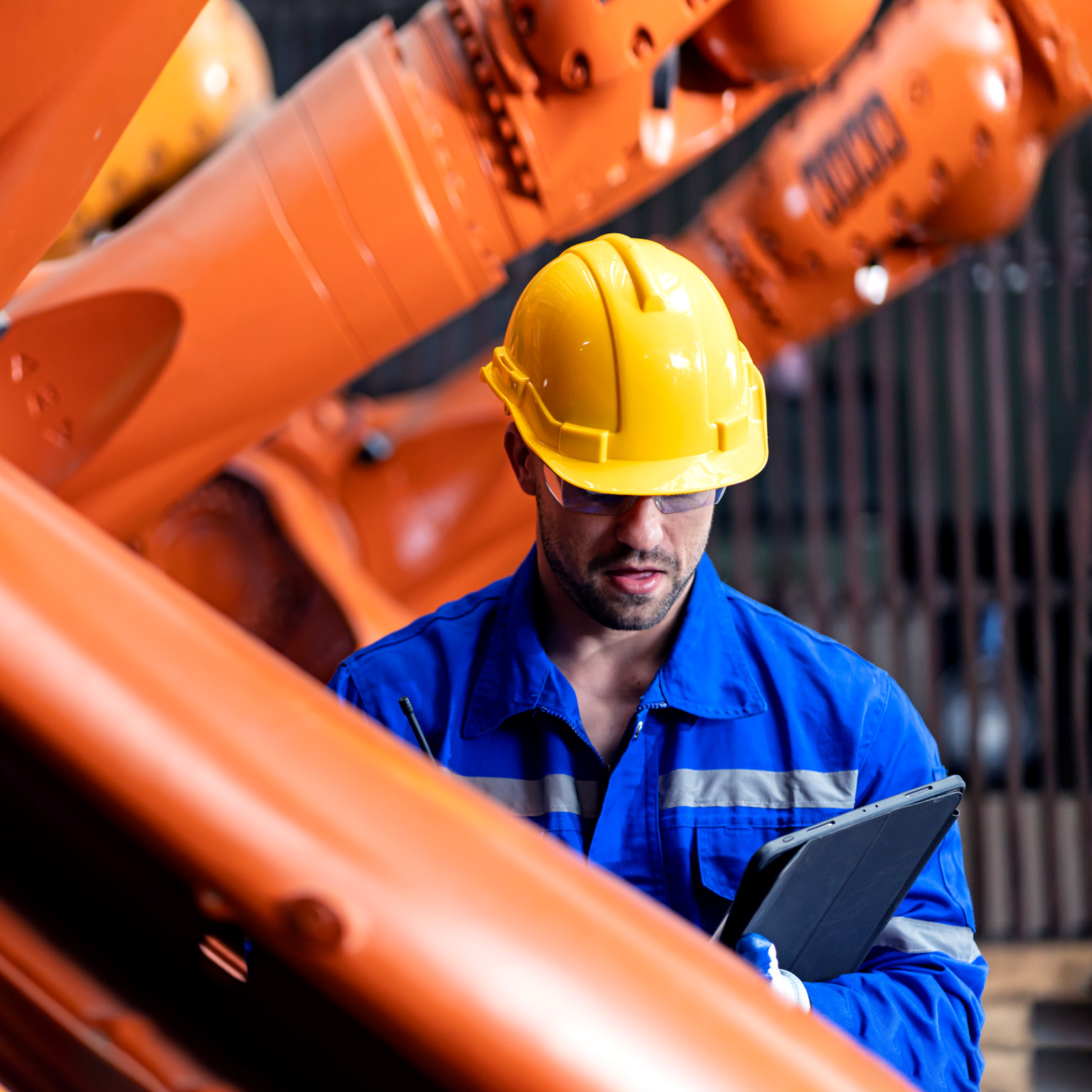 An engineer in a blue jumpsuit and yellow helmet inspects an orange steel welding robot while looking down at a tablet.