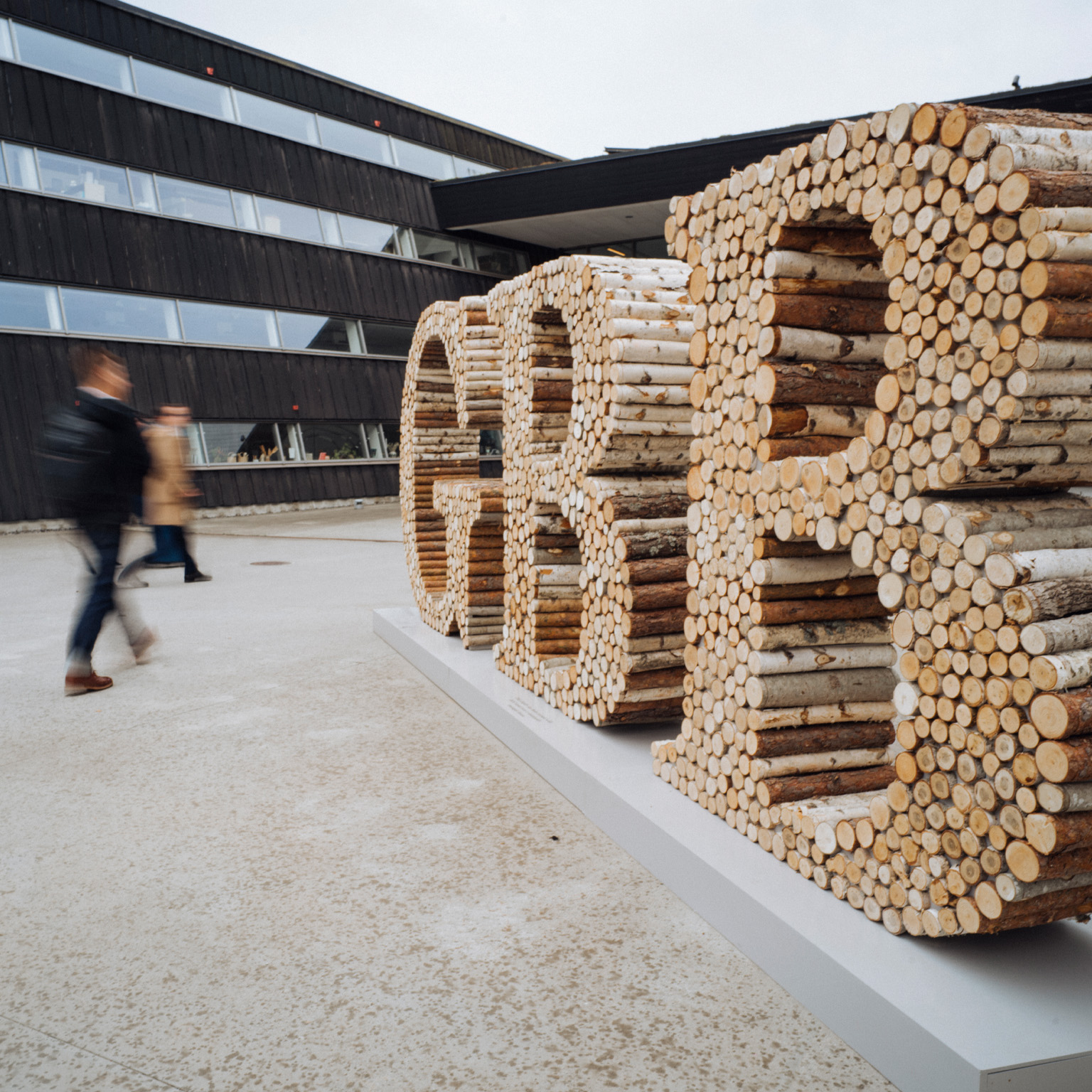 Blurred pedestrians walk past a large outdoor sculpture spelling “GBB,” constructed from stacked logs and branches, positioned in front of a modern building with dark wood cladding and long horizontal windows.