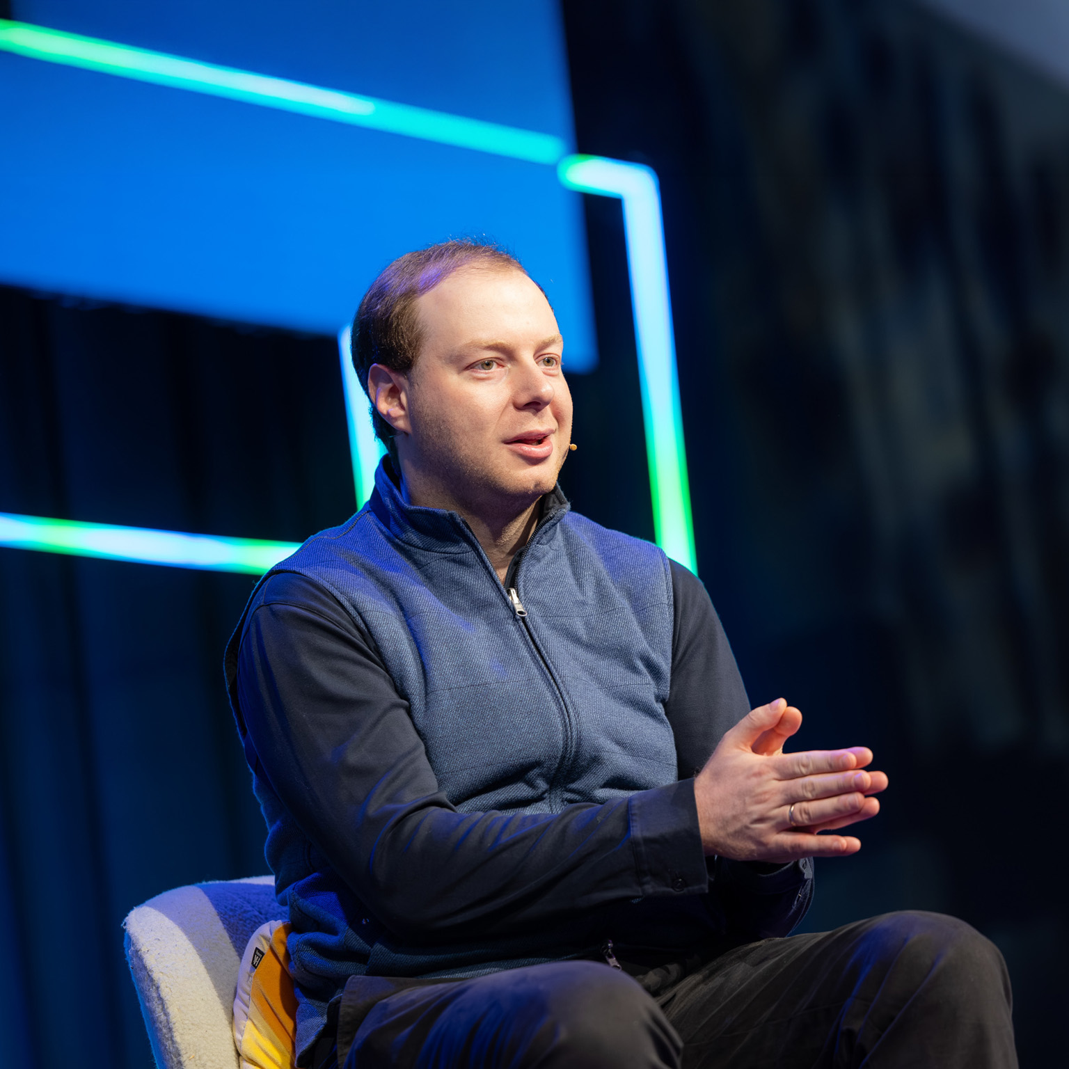 Andrey Khusid speaking on stage with a headset microphone, gesturing with his hands against a dark background accented by blue and green light strips.