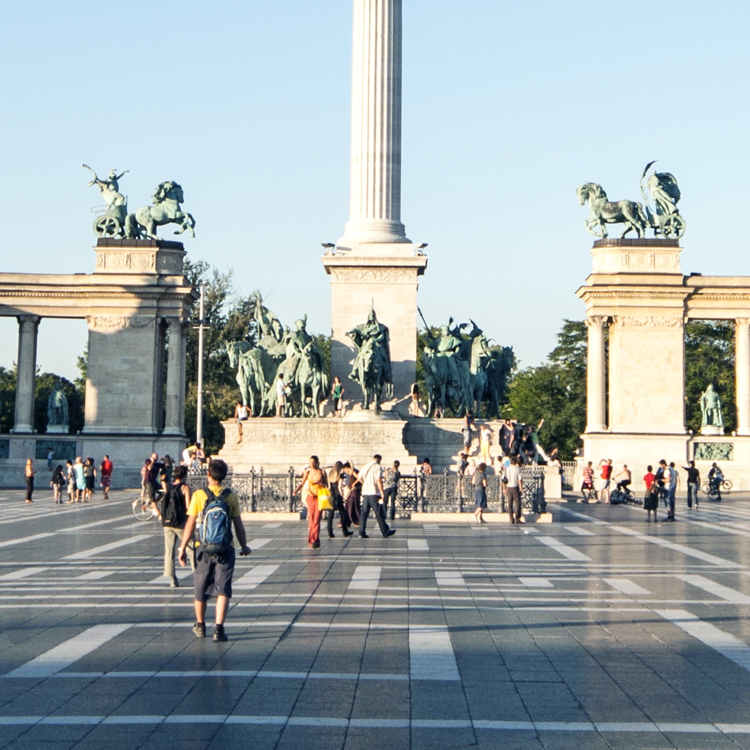 Visitors strolling and capturing images at Heroes Square in Budapest, with the Millennium column prominently displayed at the center of the view.