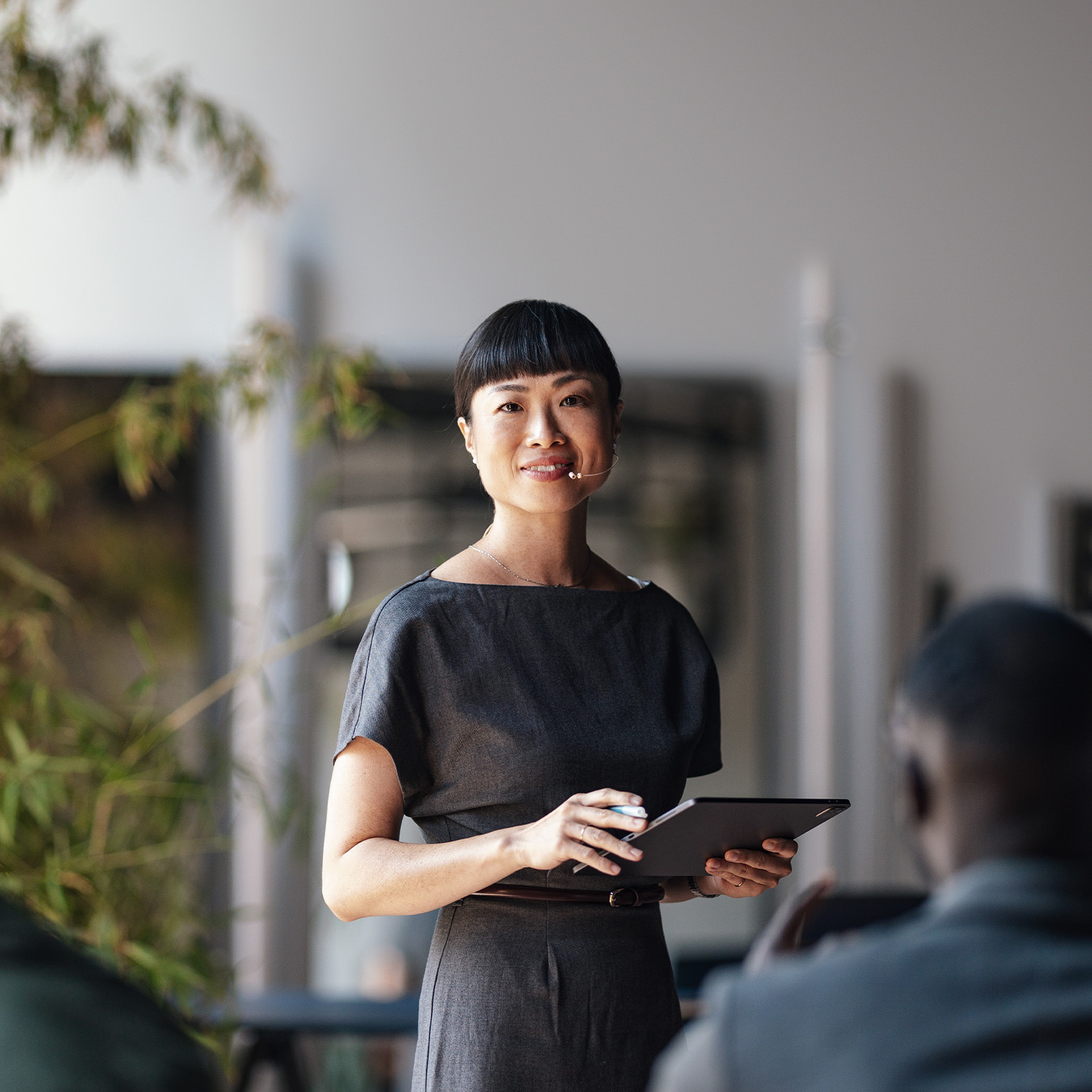 A confident presenter leads a business conference, engaging a diverse audience. The speaker is using a tablet and microphone, ensuring effective communication and interaction in a modern professional setting.