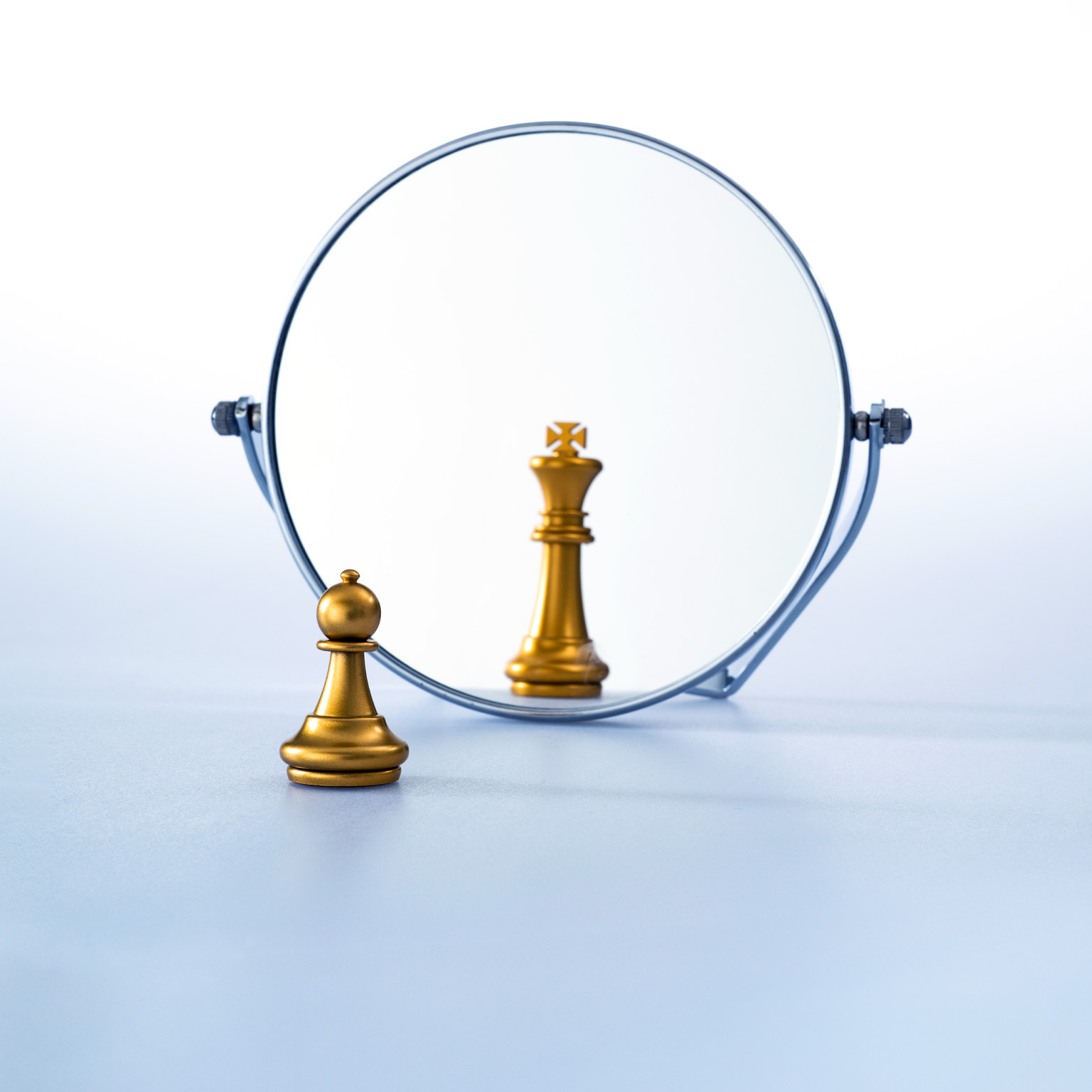 A gold pawn stands in front of a round mirror on a plain surface. In the mirror’s reflection, the pawn appears as a king piece, suggesting a shift in perception or self-image.