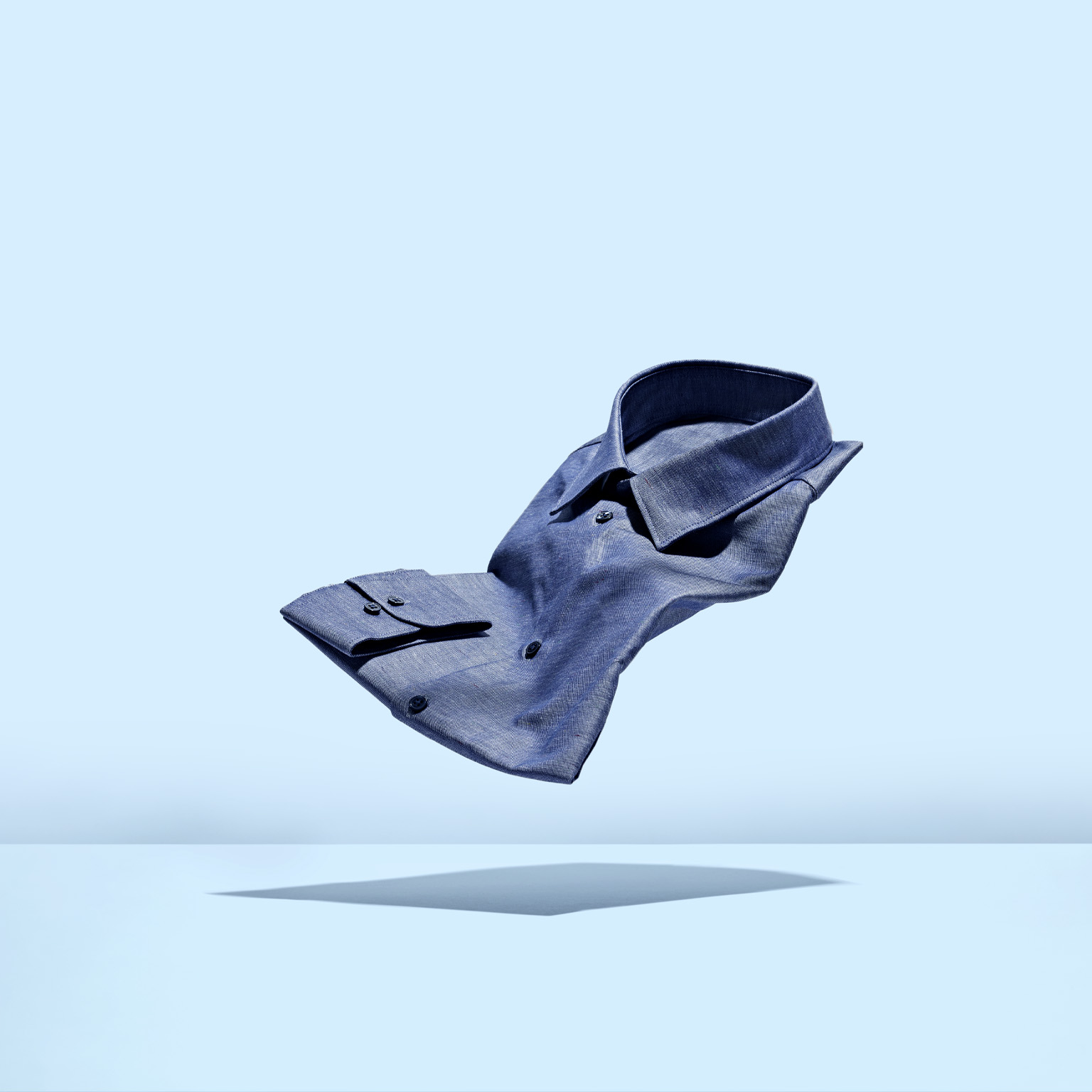 A chambray mens dress shirt floating weightlessly against a light blue background.