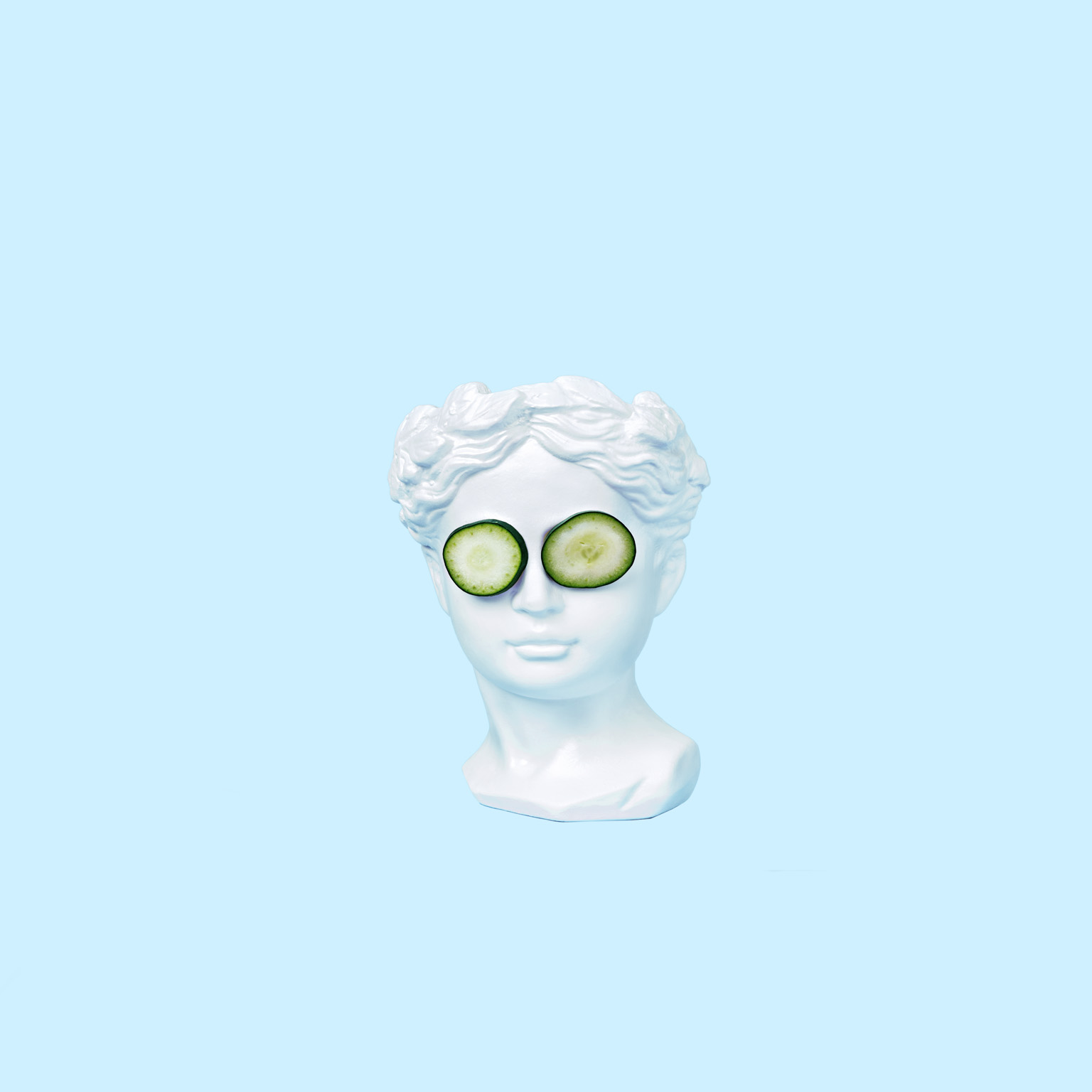 A bust of a young woman, inspired by ancient Greek art, wearing cucumber slices on her eyes and set against a light blue backdrop.