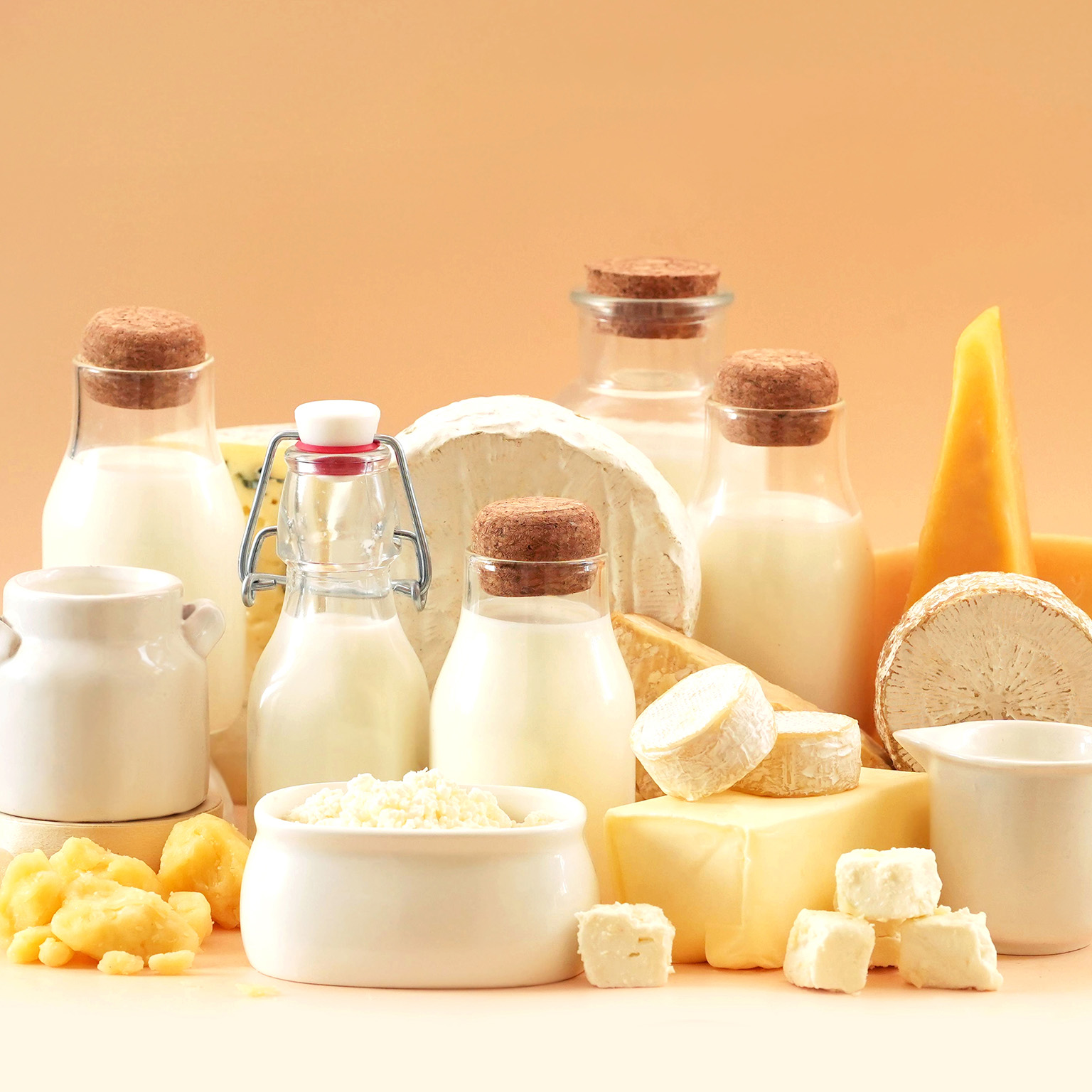 Assortment of dairy products including milk bottles, cheese varieties, butter, and cream on a light background.