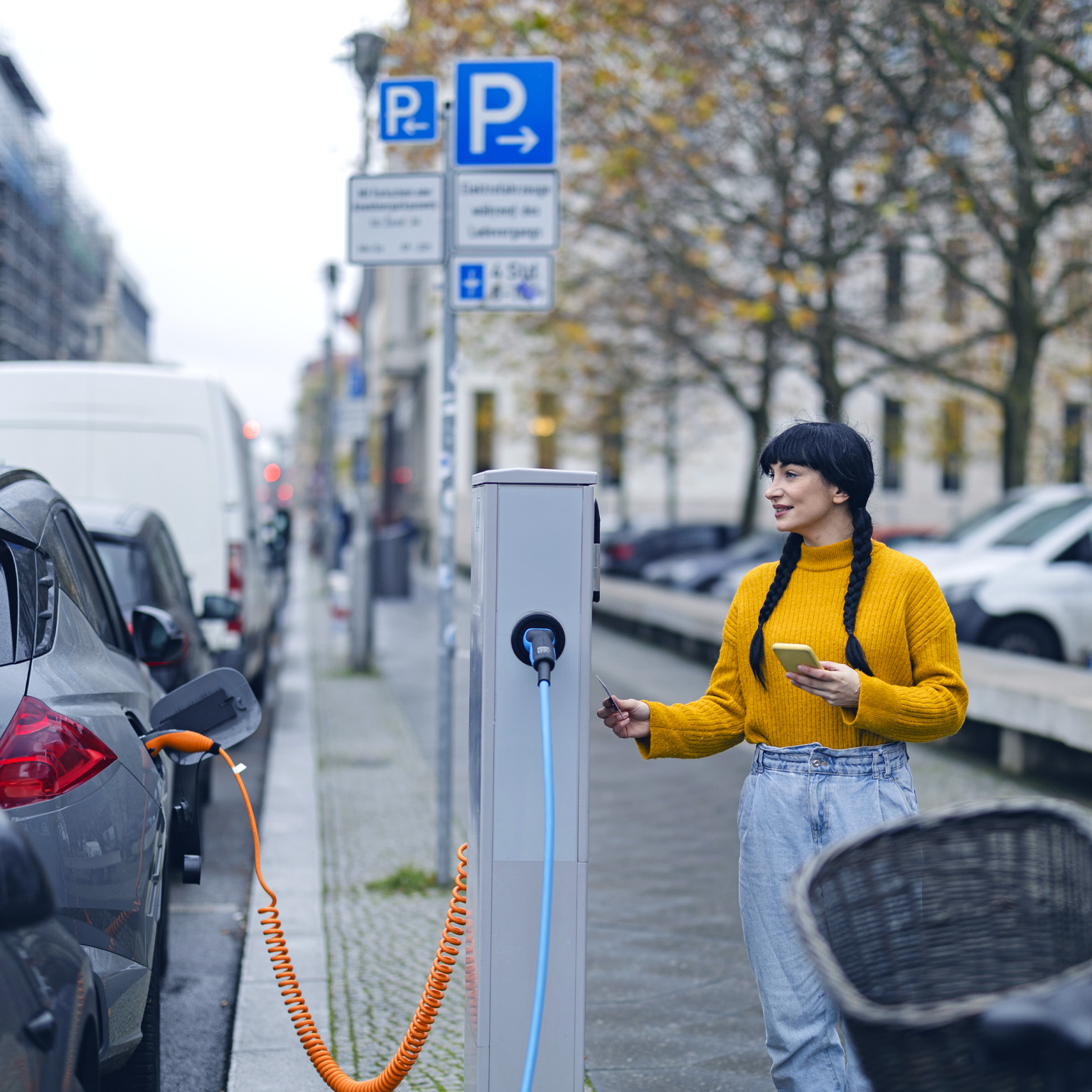 A woman with smartphone and credit card preparing to pay at an electric vehicle charging station on a tree-lined European city street.