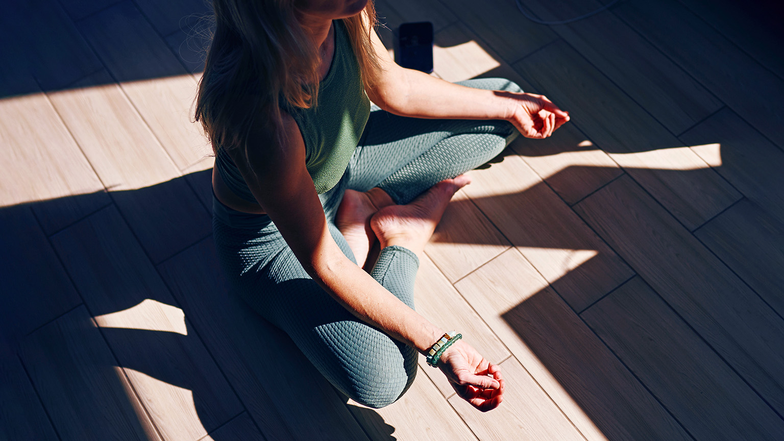 A woman sits on a wooden floor in a cross-legged meditative pose, wearing teal yoga leggings and a matching top. Sunlight streams through a window, casting shadows across the floor as she sits with her hands resting in her lap.