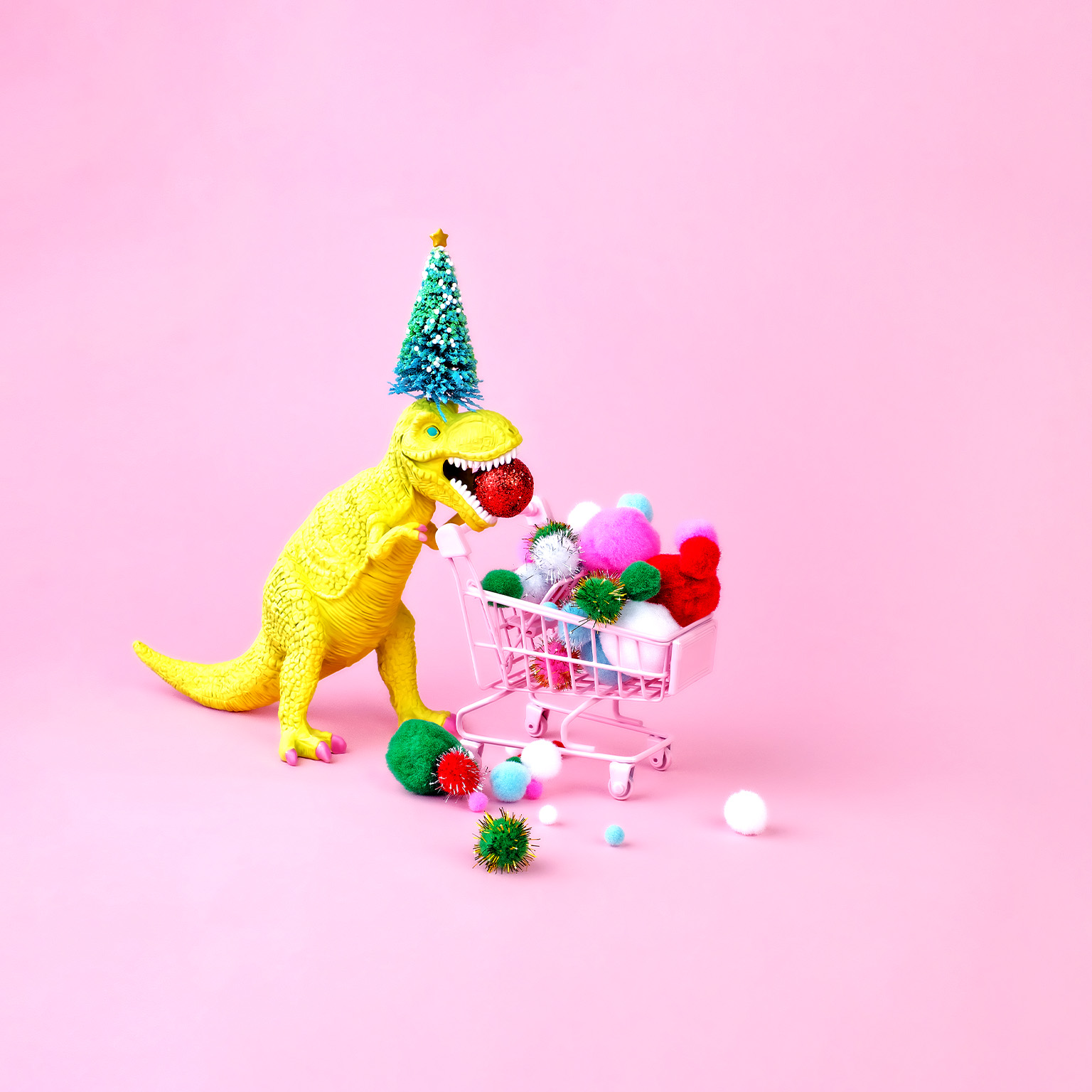 A festive image of a toy dinosaur wearing a Christmas tree on its head and filling a shopping cart with multicolored puff balls.