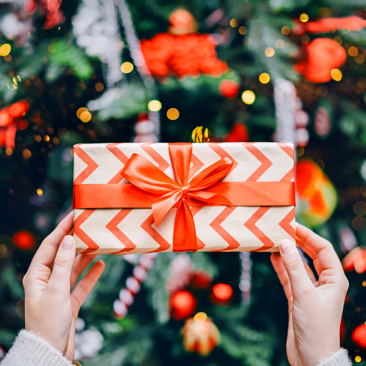 A pair of hands holding a wrapped gift box decorated with a red chevron pattern and tied with a bright red ribbon. In the background, colorful lights and ornaments create a festive, holiday atmosphere.