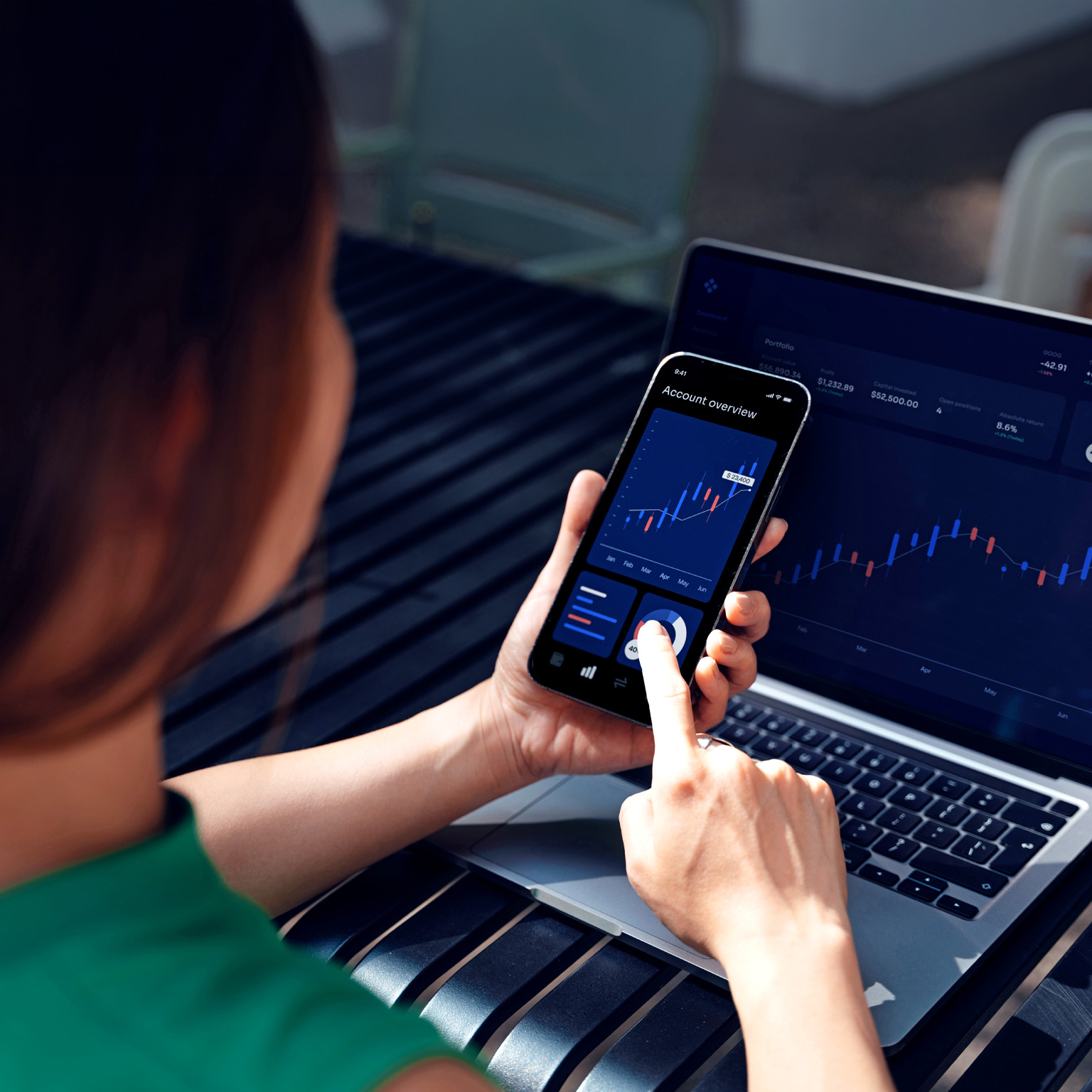 A person uses a smartphone displaying financial charts while viewing a matching investment dashboard on a laptop.