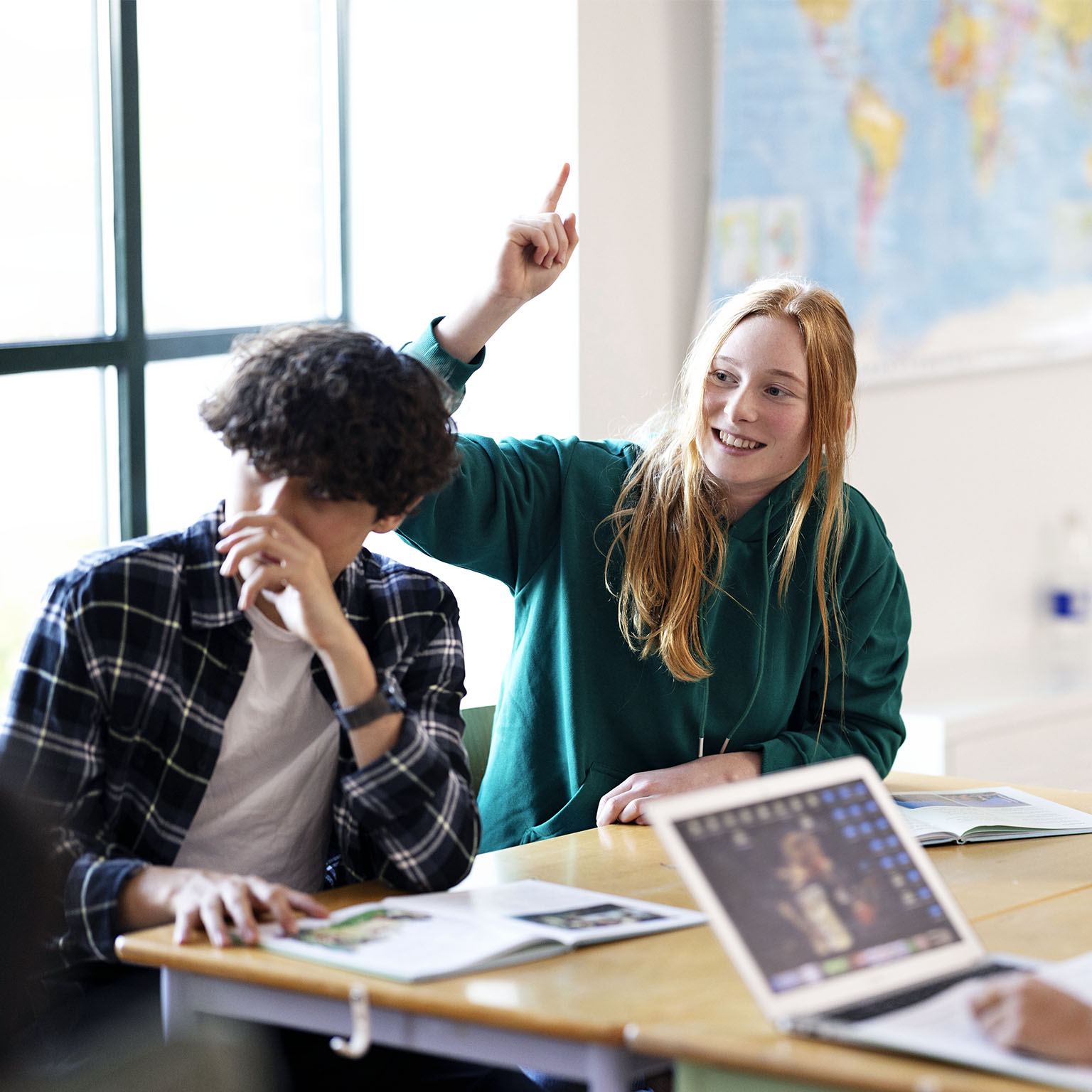 A group of students sitting at a classroom table, with a red-haired student in a green hoodie smiling and raising her hand while others, including one with a laptop, also participate.