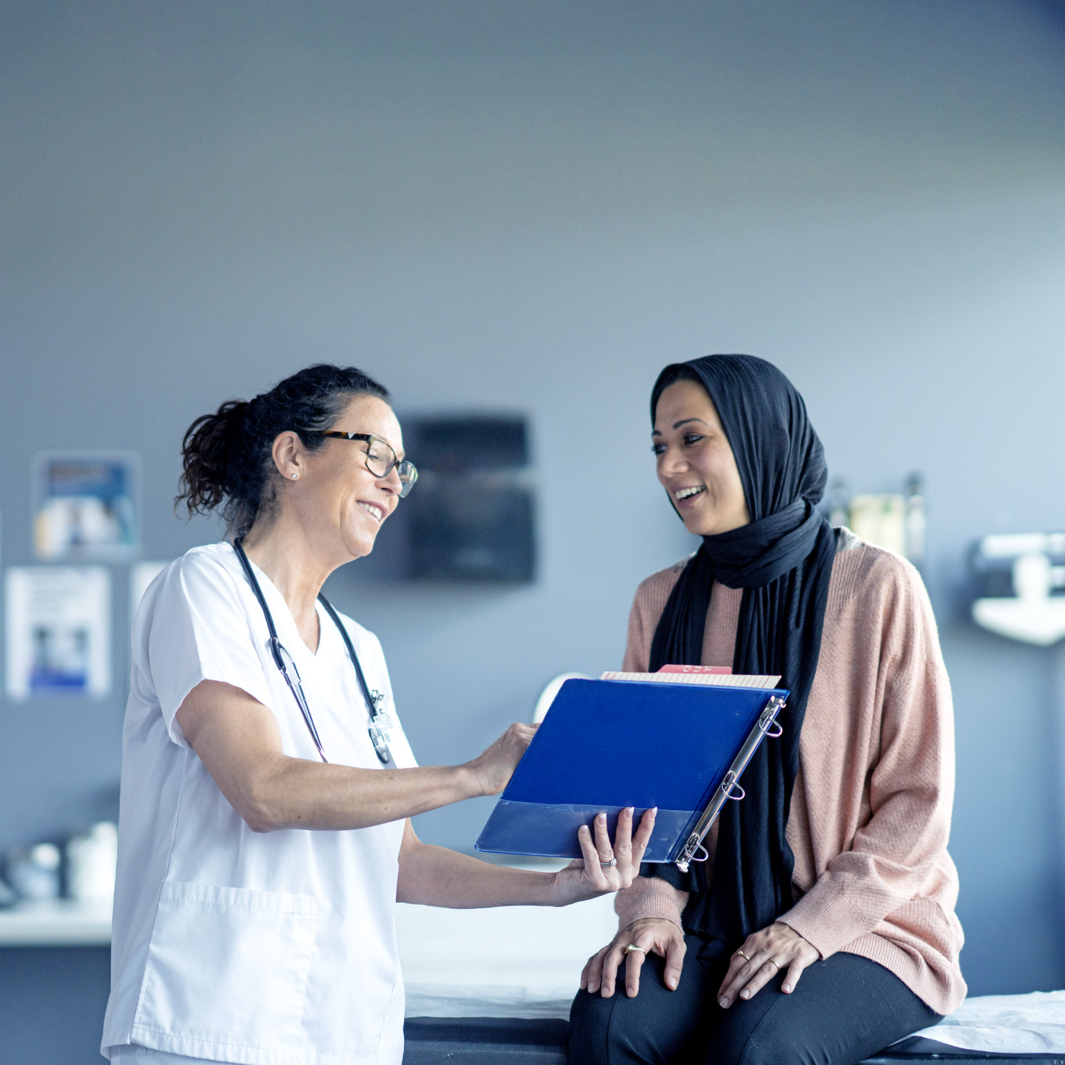 In an examination room with medical equipment behind them, a female patient wearing a black hijab and a pink sweater is greeted by a female doctor dressed in a white coat and stethoscope. The doctor holds a clipboard and smiles warmly at the patient.