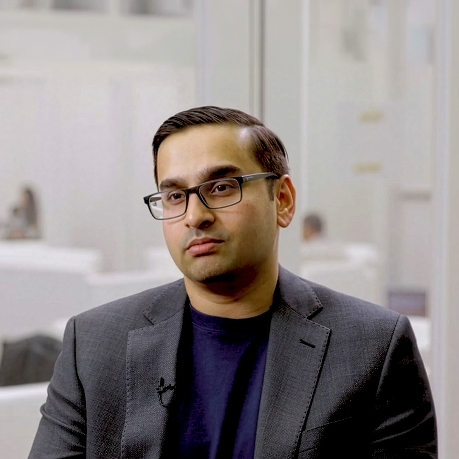 Ani Kelker wearing glasses and a dark blazer seated in a modern office during an interview. 