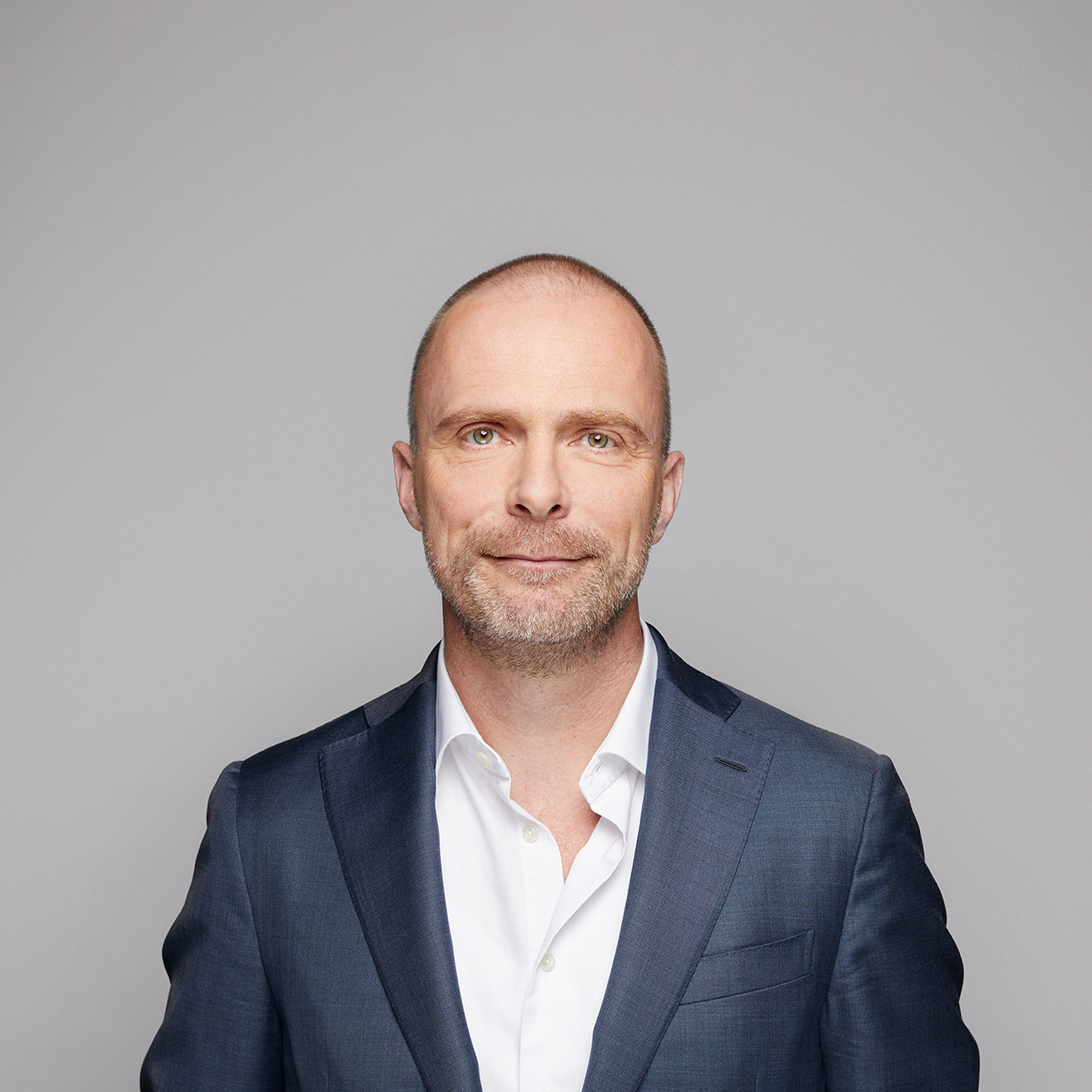 A portrait of Robert Carsouw, with a light beard, and a easy smile. He is wearing a dark blue suit and a white, open-collared shirt against a light gray background.