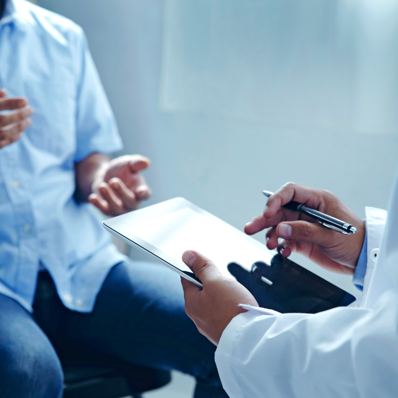 A close up of a doctor's hands holding a tablet while talking to a patient about his treatment plan.