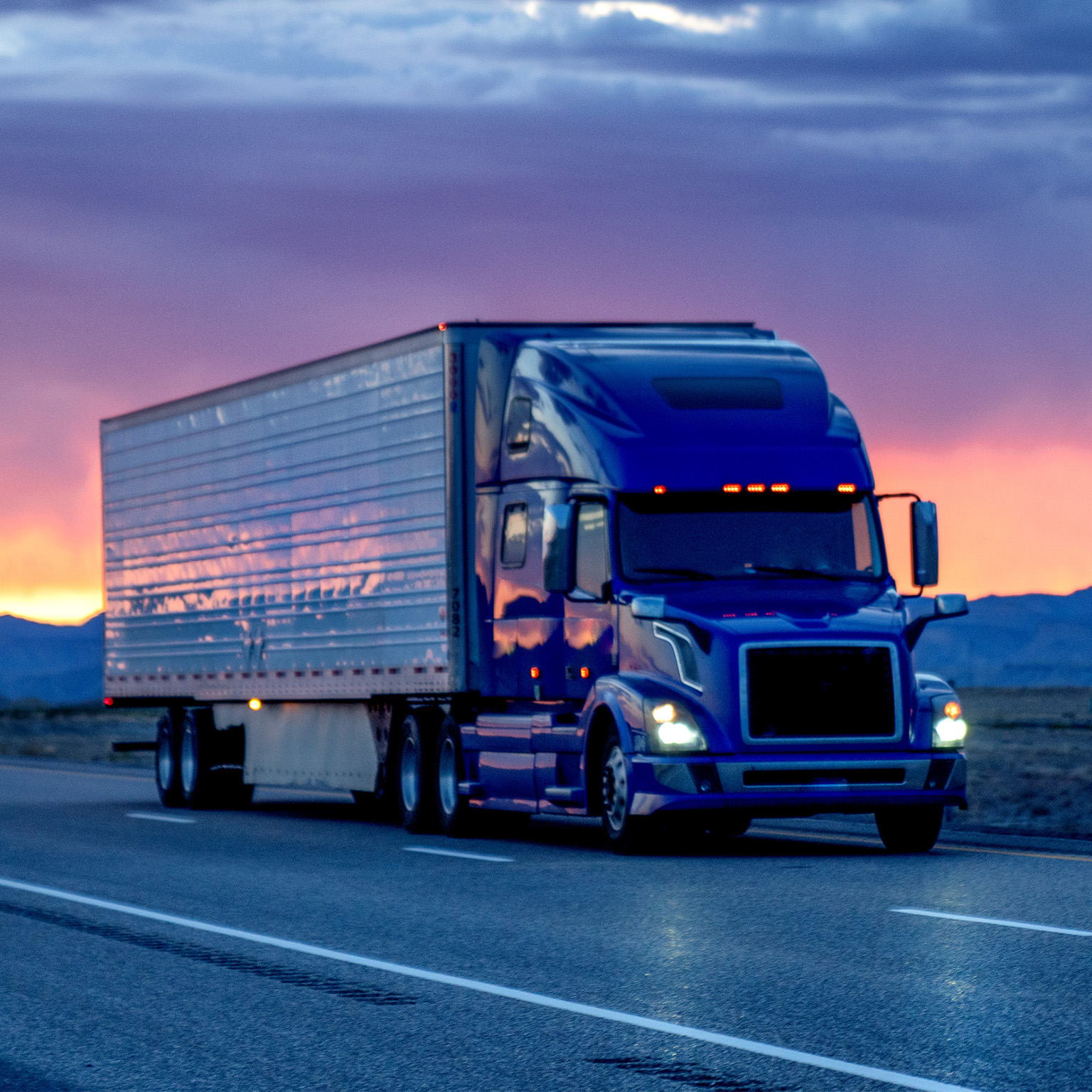 Blue semi-truck driving on a highway at sunset with cloudy sky in the background
