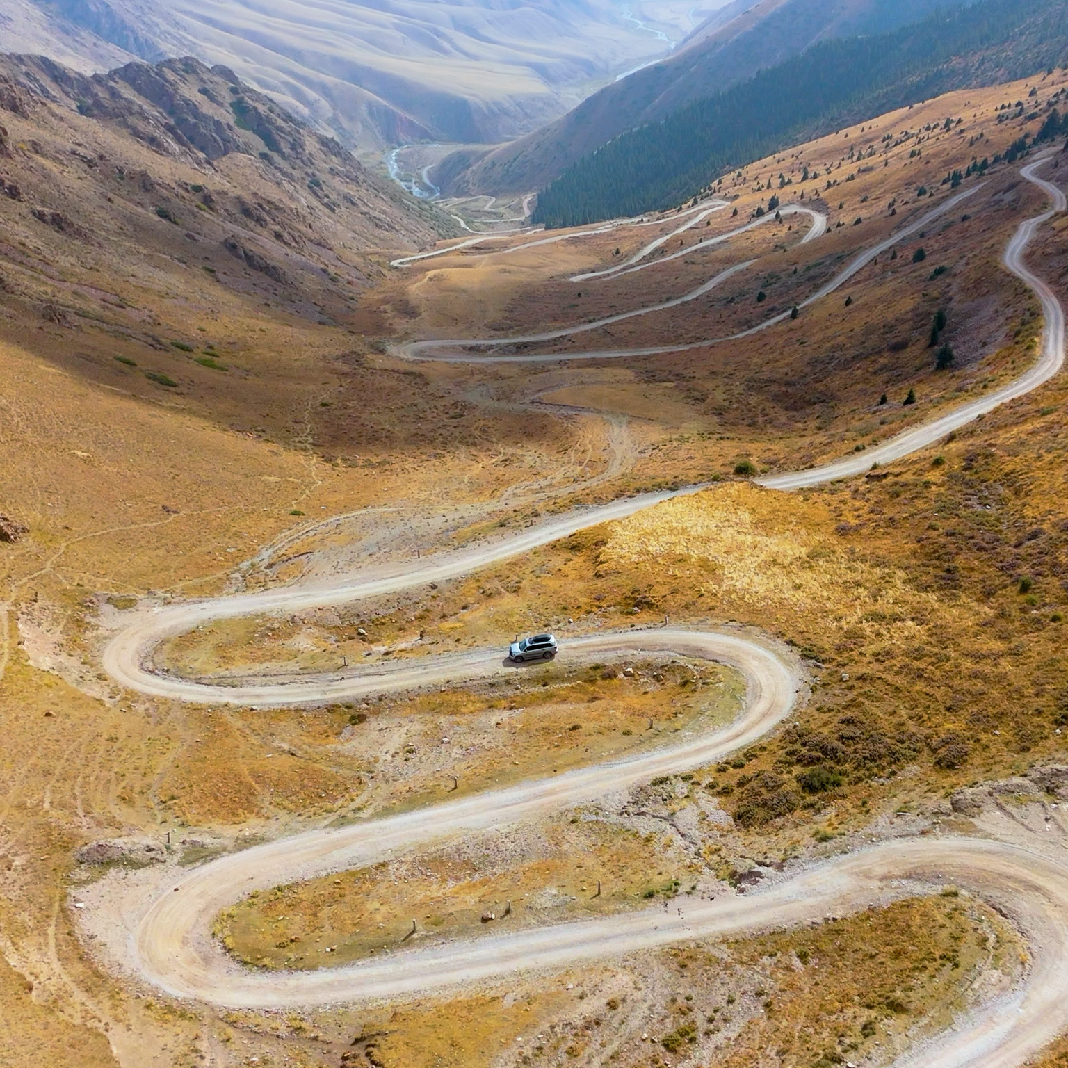 A silver SUV makes navigates a winding dirt road as it snakes through a wide mountain valley, cutting sharp switchbacks across dry, golden-brown slopes. Steep ridges rise on both sides, with sparse vegetation and distant peaks fading into a hazy, expansive backdrop.