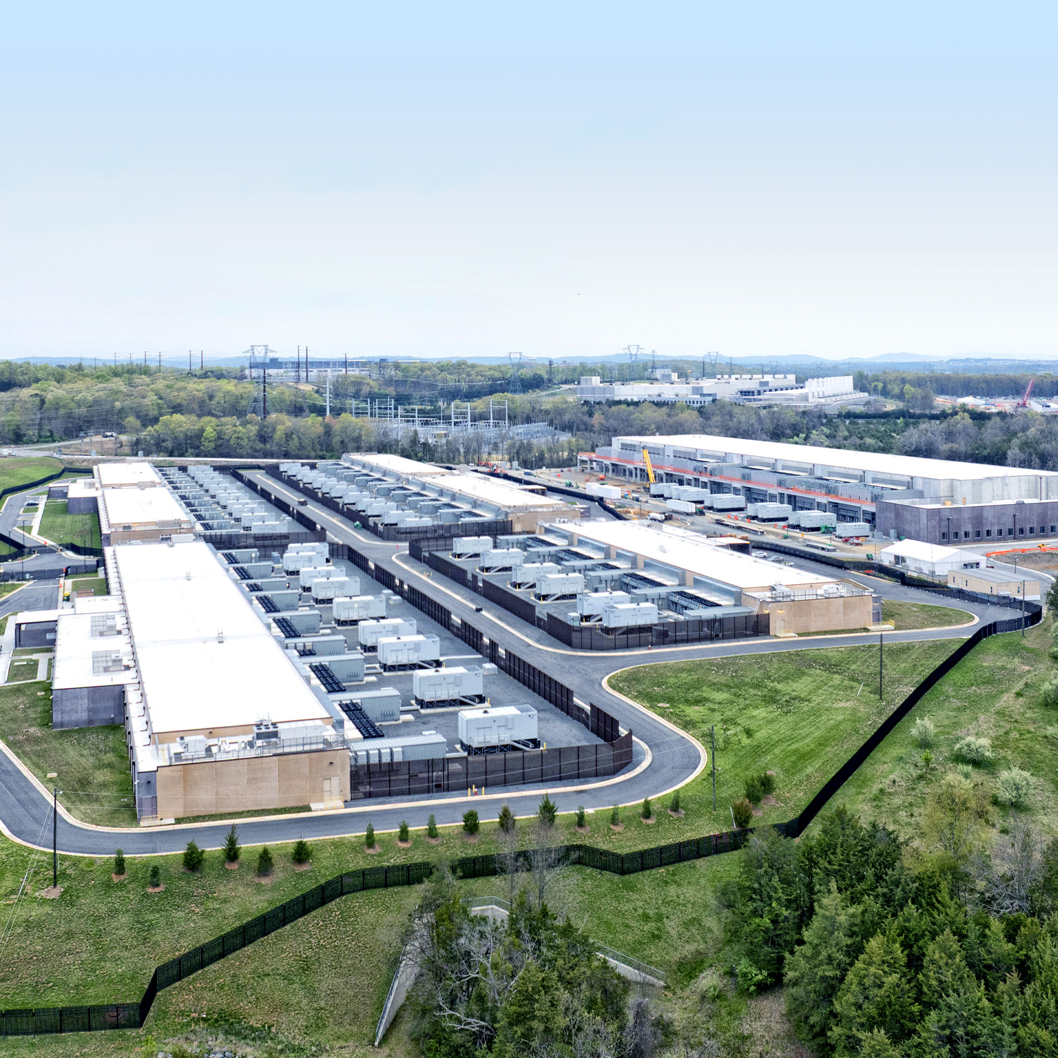 A sprawling data center complex surrounded by greenery and infrastructure. Several rectangular buildings with white roofs are visible, densely packed with cooling units or similar equipment, with at least one building under construction.