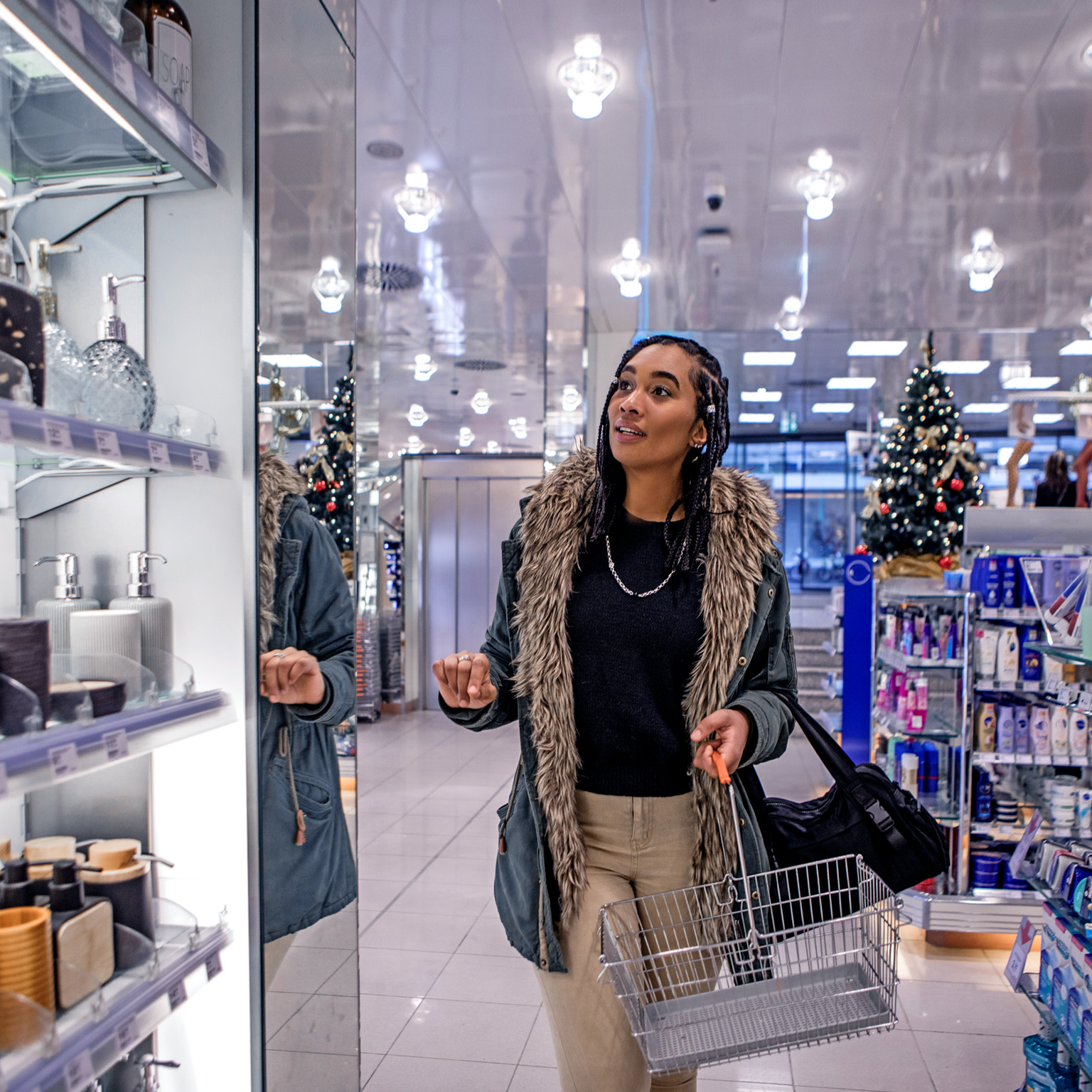 A woman walking down an aisle in a store while looking at personal care items. She is wearing a warm coat and holding a shopping basket, store holiday decorations can be seen in the background.