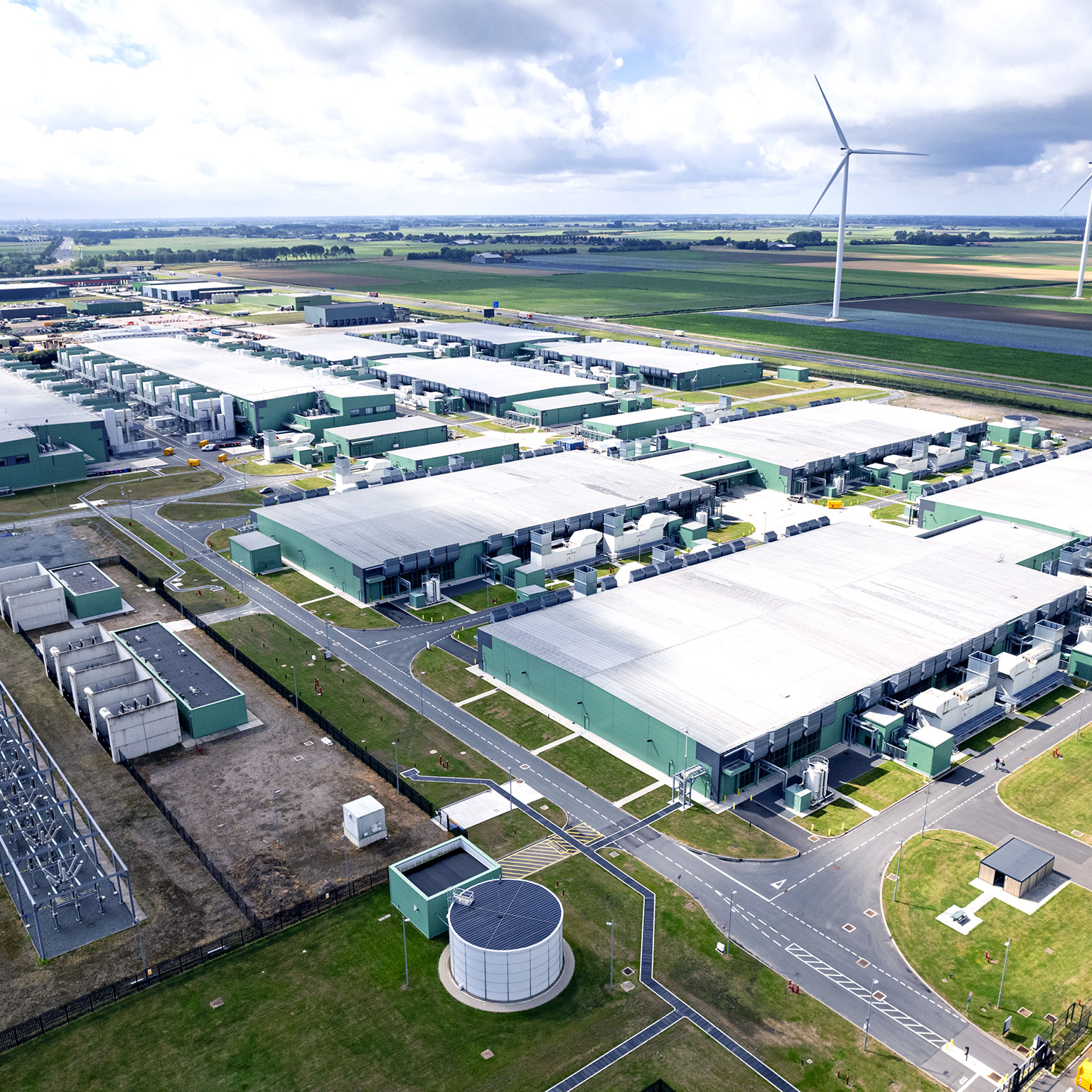 High angle view of a data center surrounded by green fields and wind turbines on the horizon. 